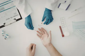 Medical professional drawing blood from a patient's finger for a COVID-19 test; test materials on white table.