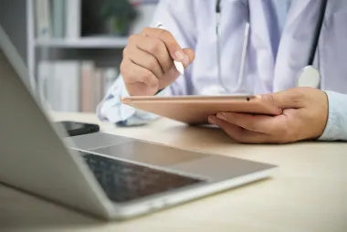 Doctor in white coat using a tablet and laptop at a desk.