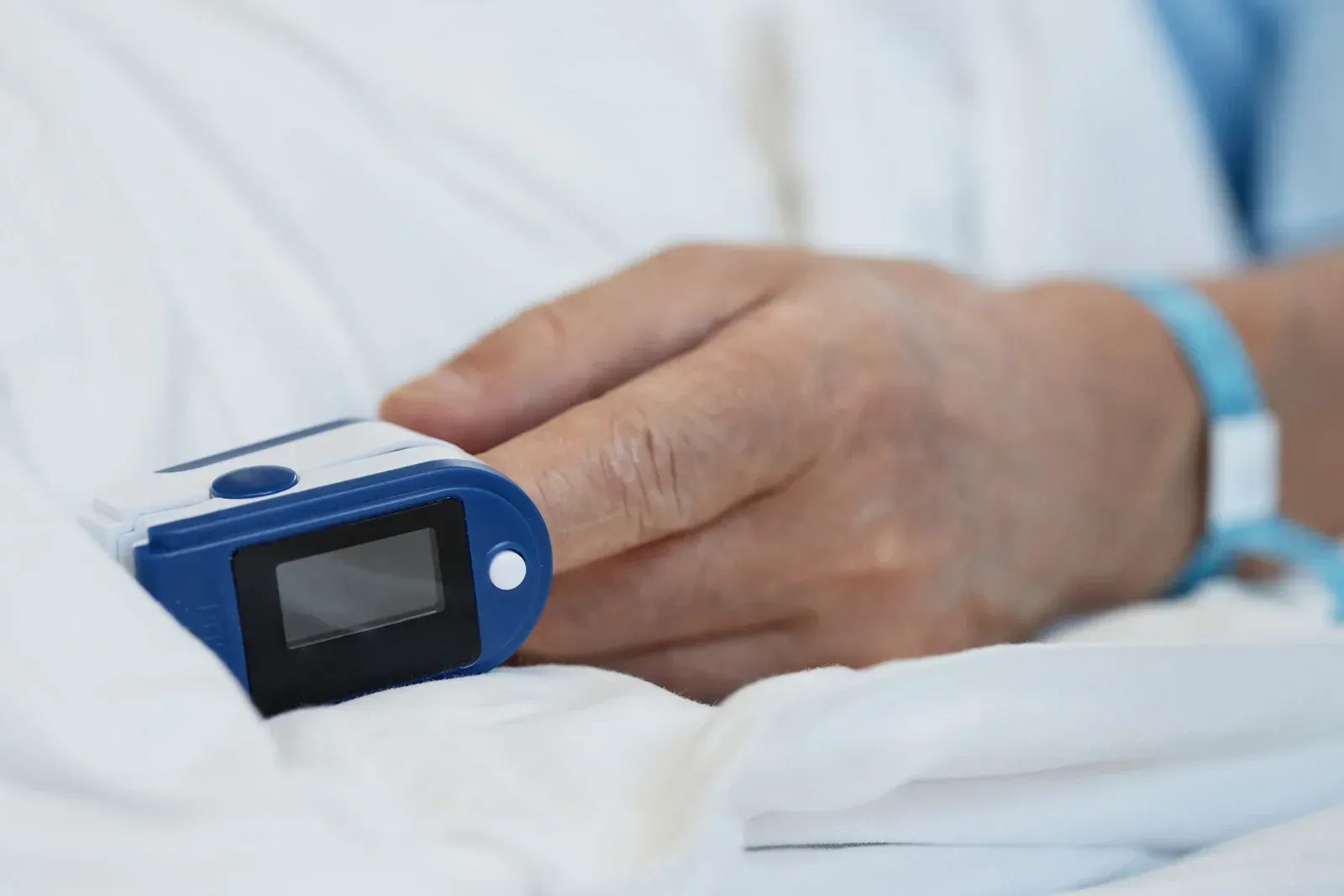 Hand with finger in a pulse oximeter, resting on a white sheet.