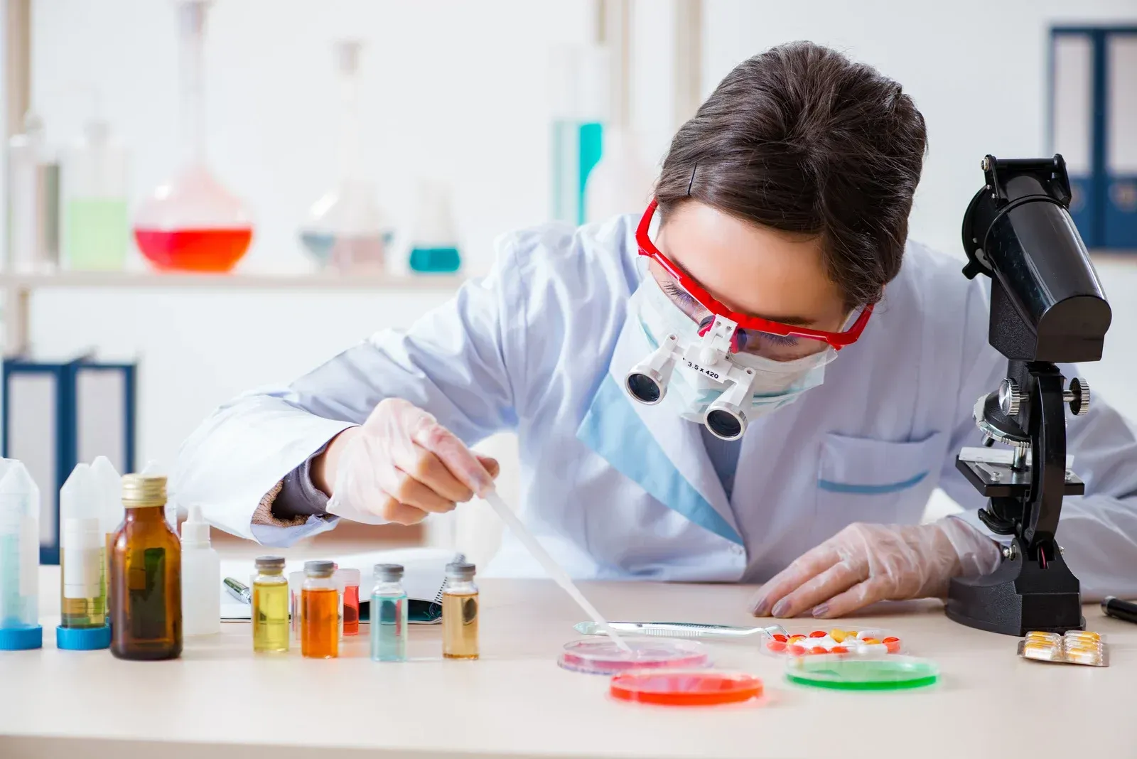 Scientist in lab coat uses pipette to examine petri dishes with colorful samples under a microscope.