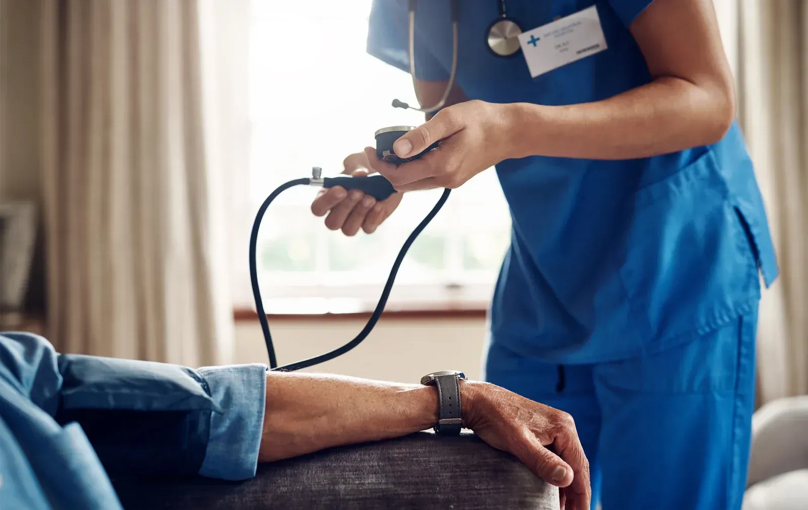 Nurse taking a patient's blood pressure with a stethoscope in a home setting.