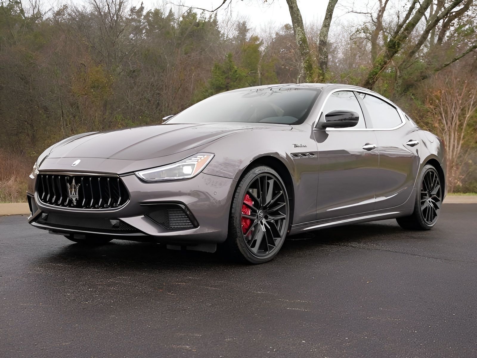 Gray Maserati Ghibli sedan parked on wet pavement; black grille, red brake calipers.