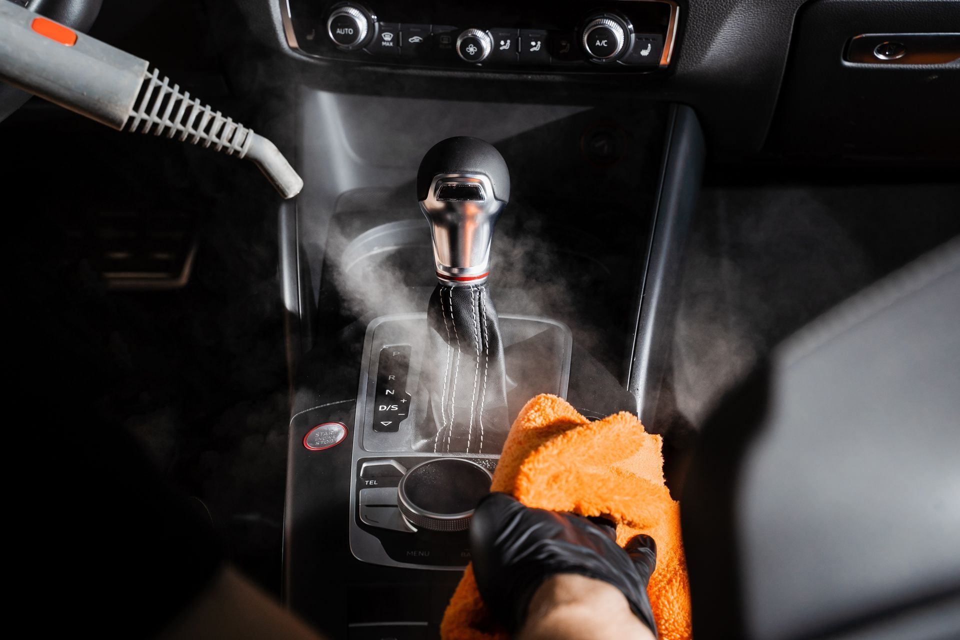 A person steam-cleaning a car's center console with an orange microfiber cloth and black gloves.