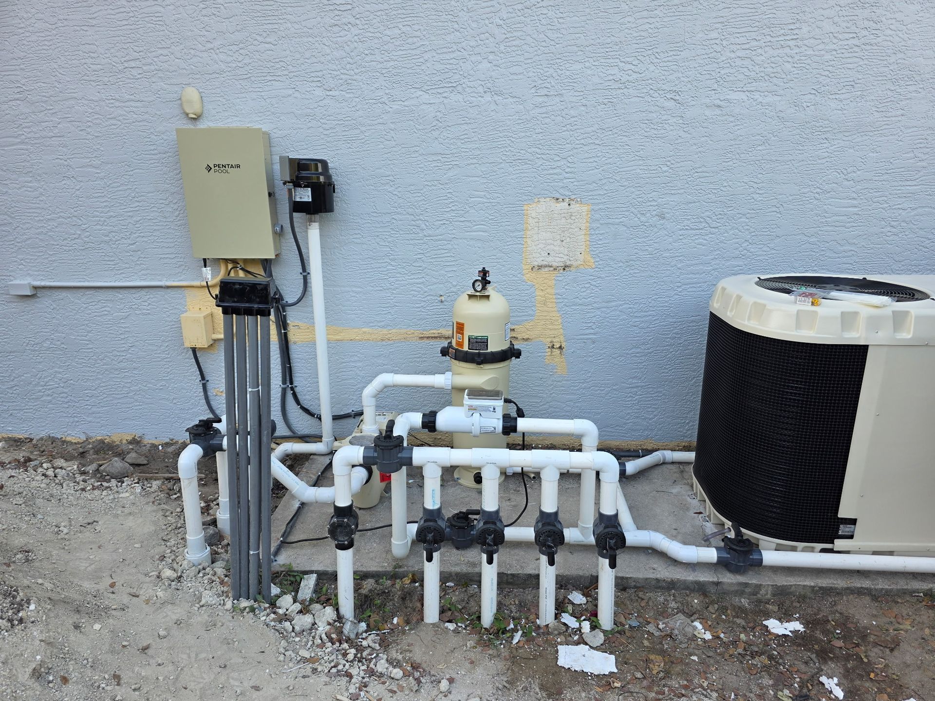 Outdoor HVAC and water piping equipment beside a stucco wall, with a beige control box and metal pipes
