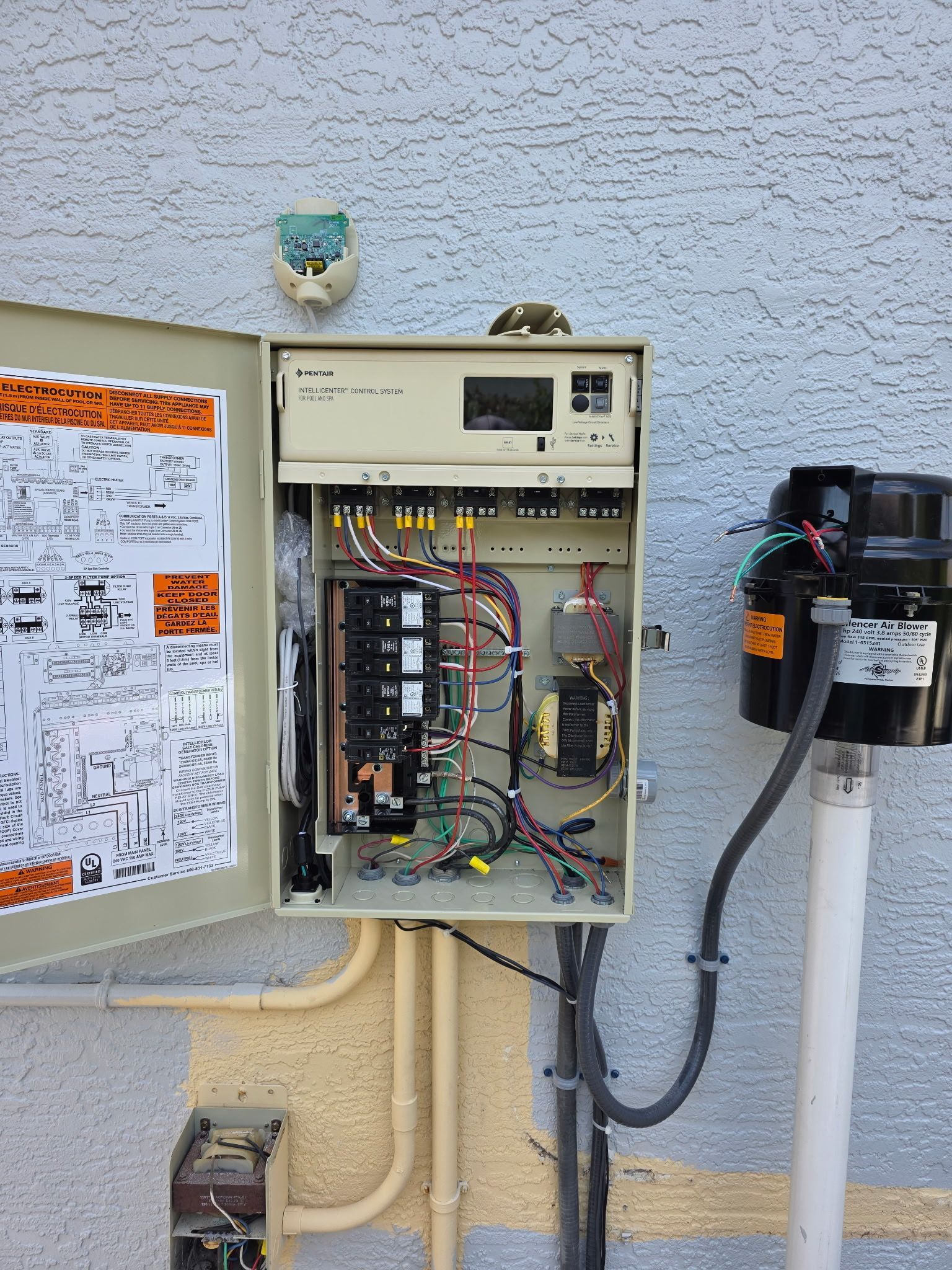 Open electrical panel with exposed wiring beside a wall-mounted utility meter box and nearby notice board.