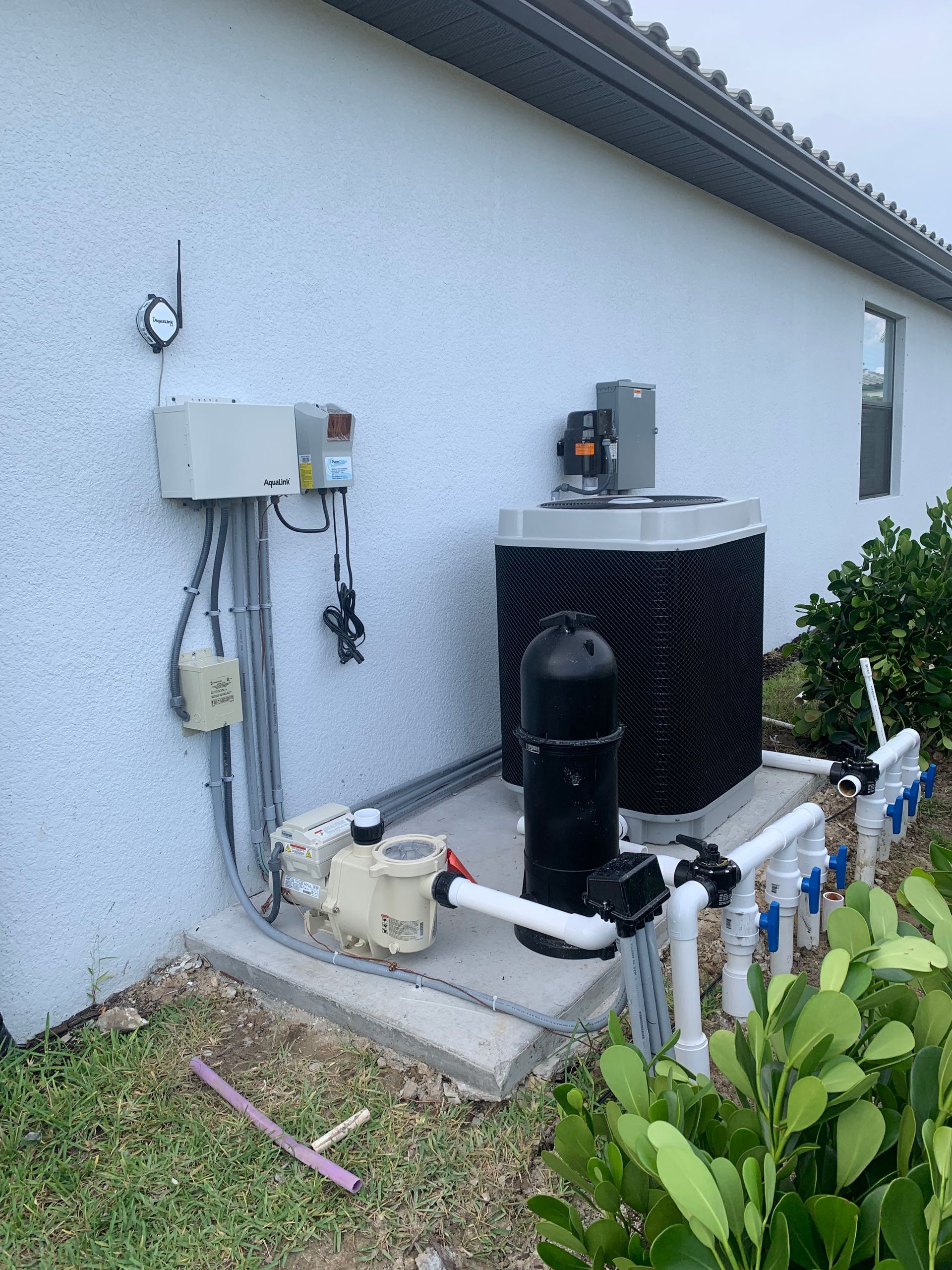 Outdoor heat pump and utility equipment mounted beside a white house wall with pipes and shrubs nearby