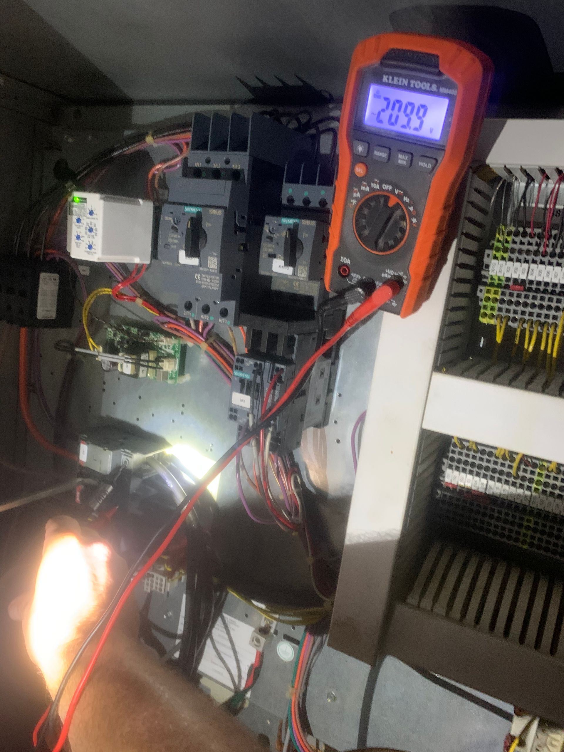 Orange multimeter clipped to electrical control panel with wiring and bright light source below