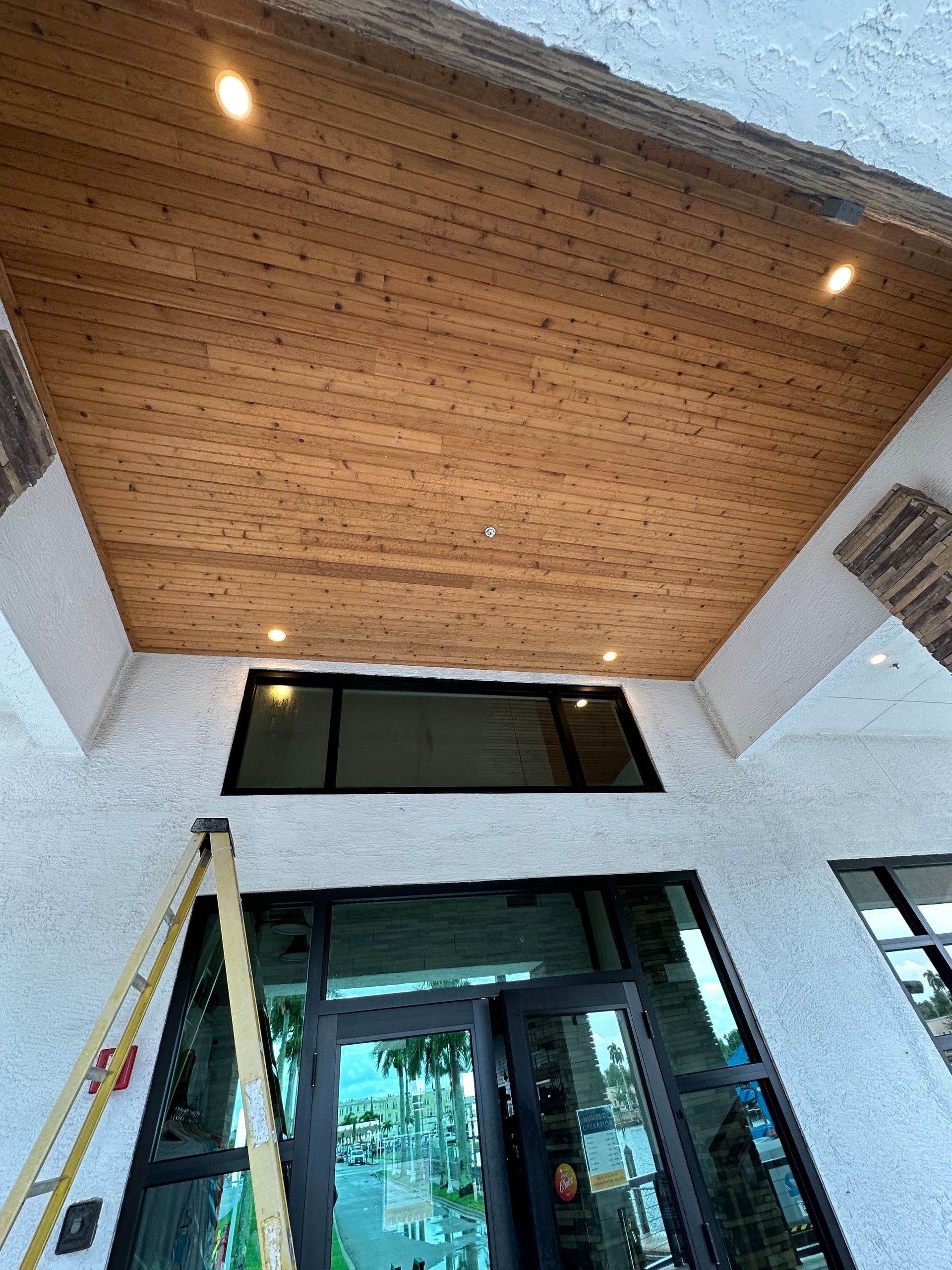 Front entrance with glass doors, ladder, and a wood-paneled ceiling with recessed lights.