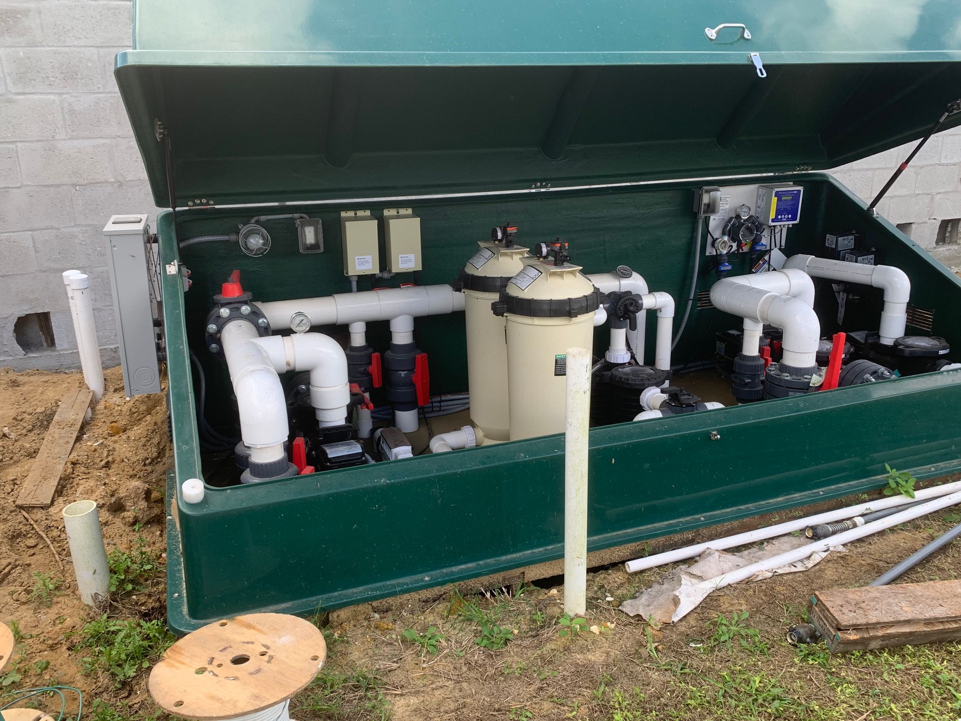 Open green utility enclosure with pipes, valves, and a beige tank beside a building outside