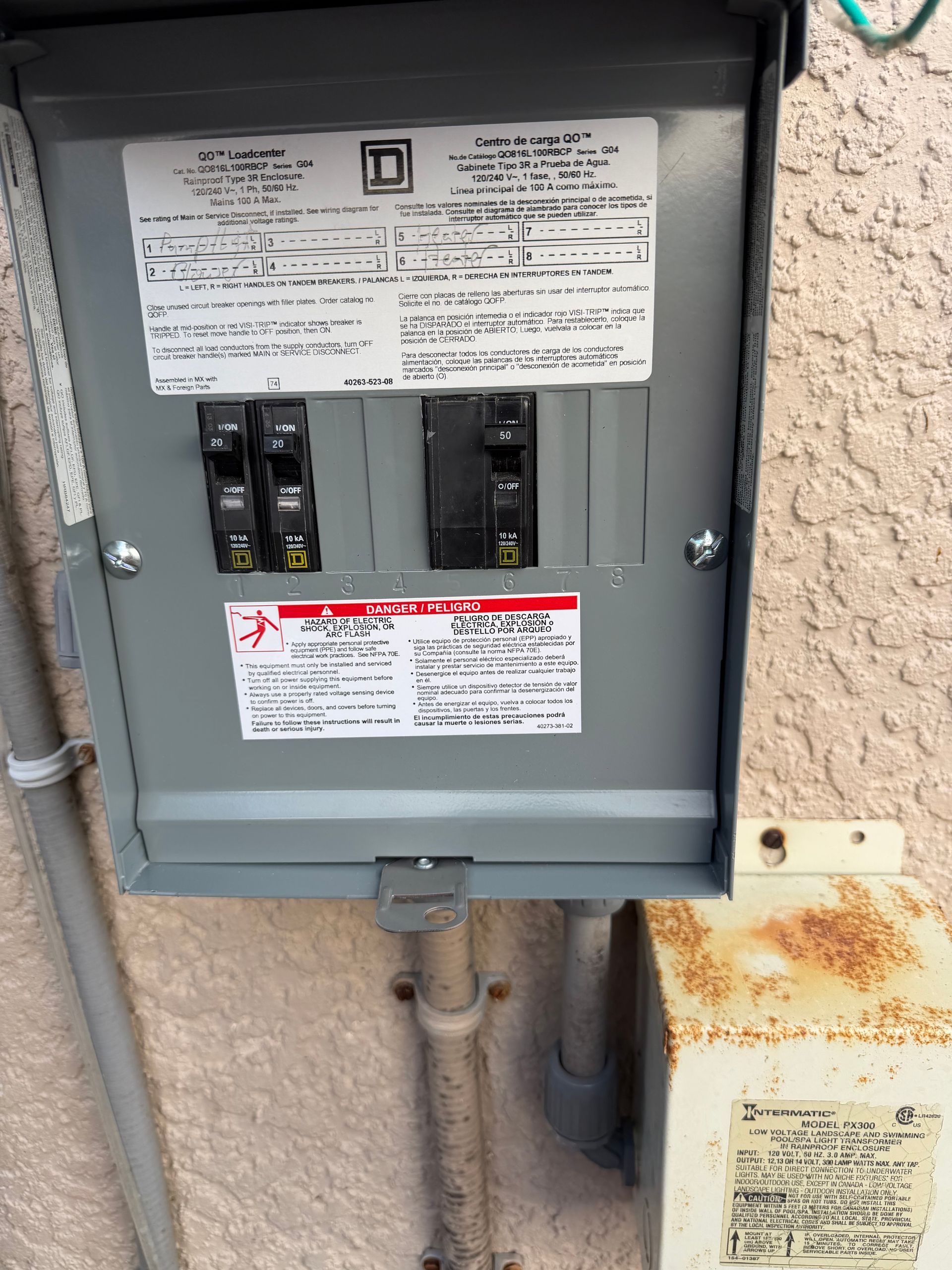 Open electrical breaker panel on a stucco wall with warning labels and rusted conduit below