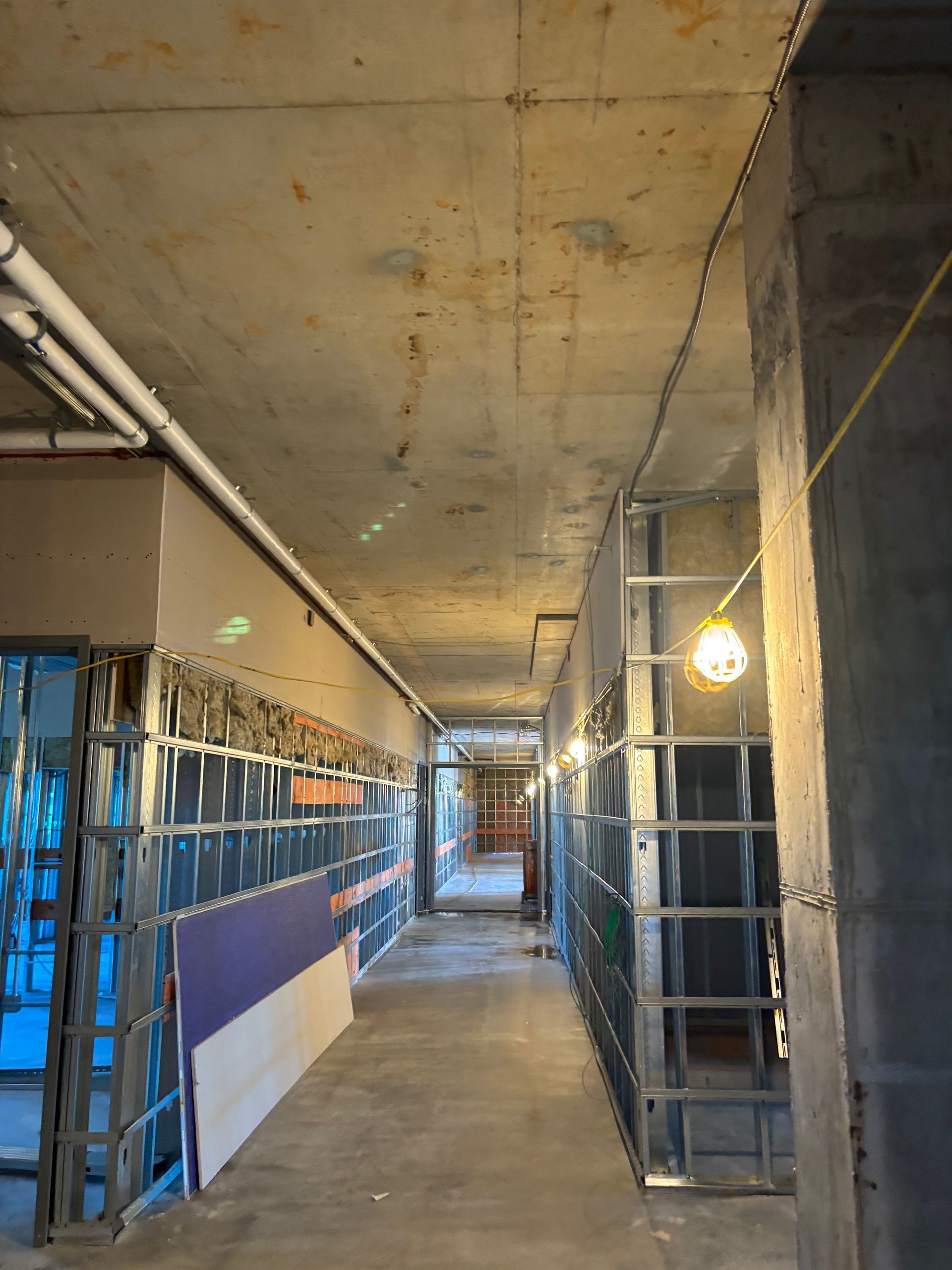 Unfinished hallway under construction with exposed ceiling, scaffolding, and blue protective sheeting along the walls