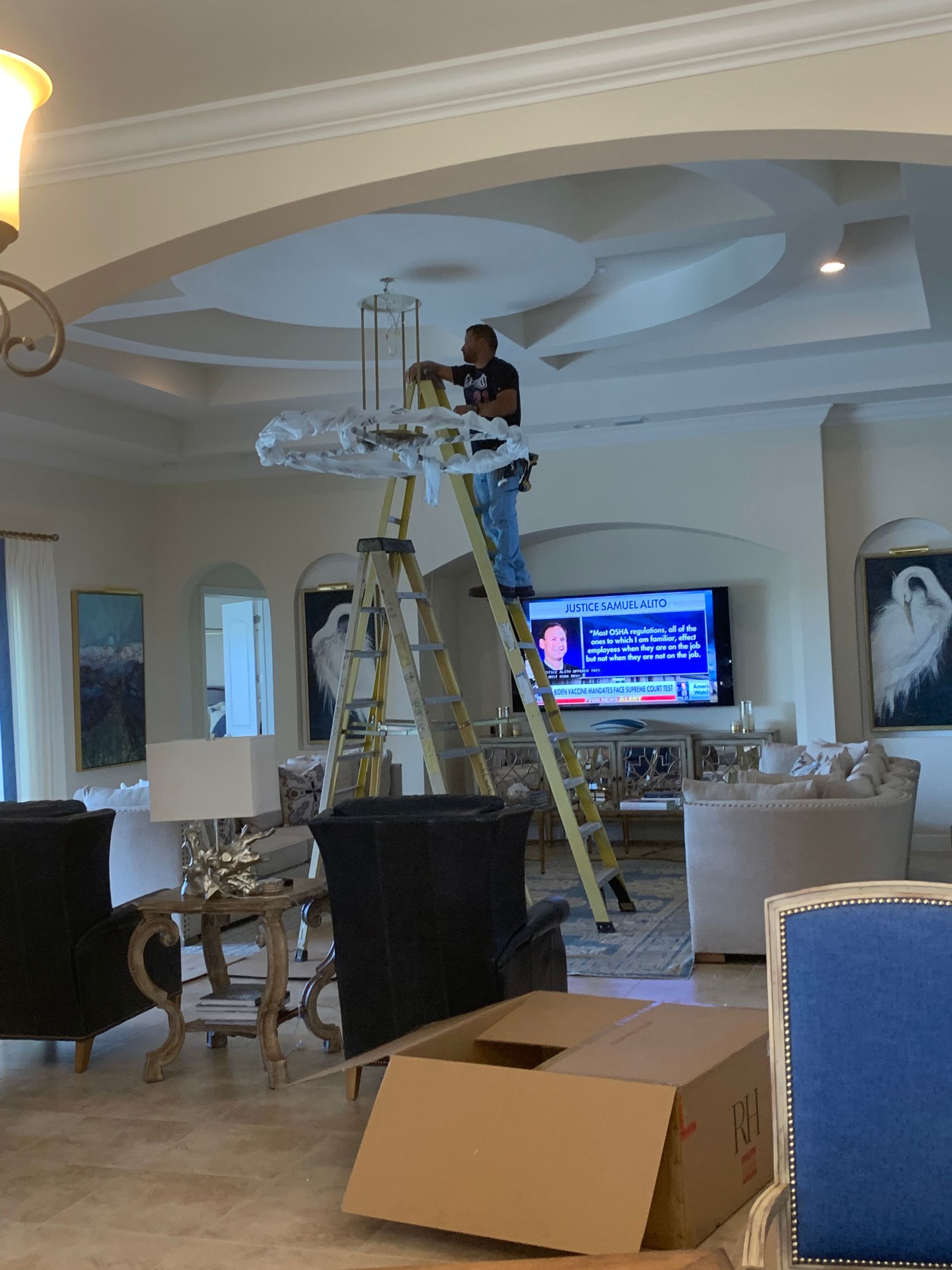 Person on ladder installing a chandelier in a living room, with cardboard box and TV nearby.