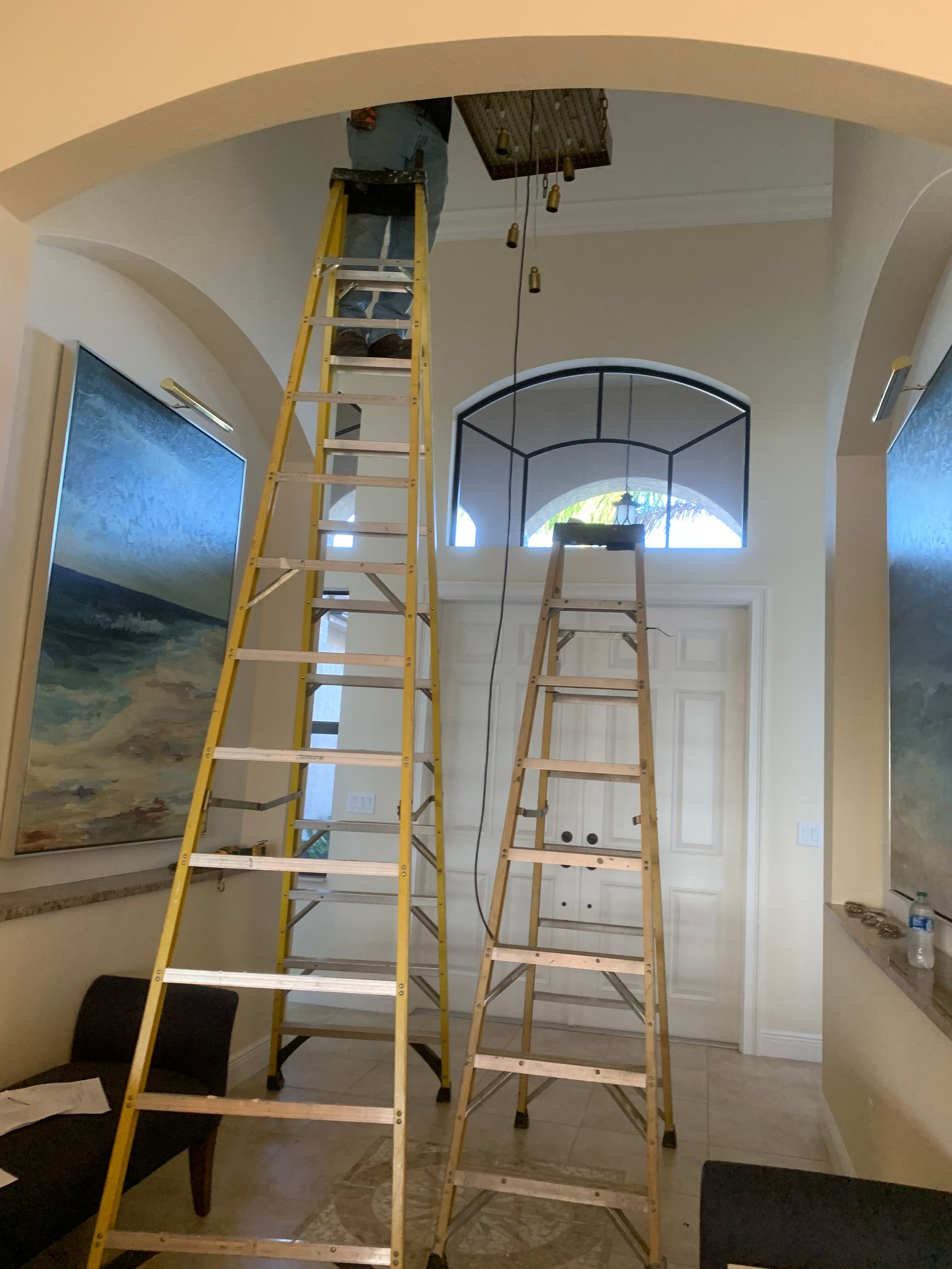 Two ladders in a hallway beneath a ceiling opening, with a painting on the left wall.