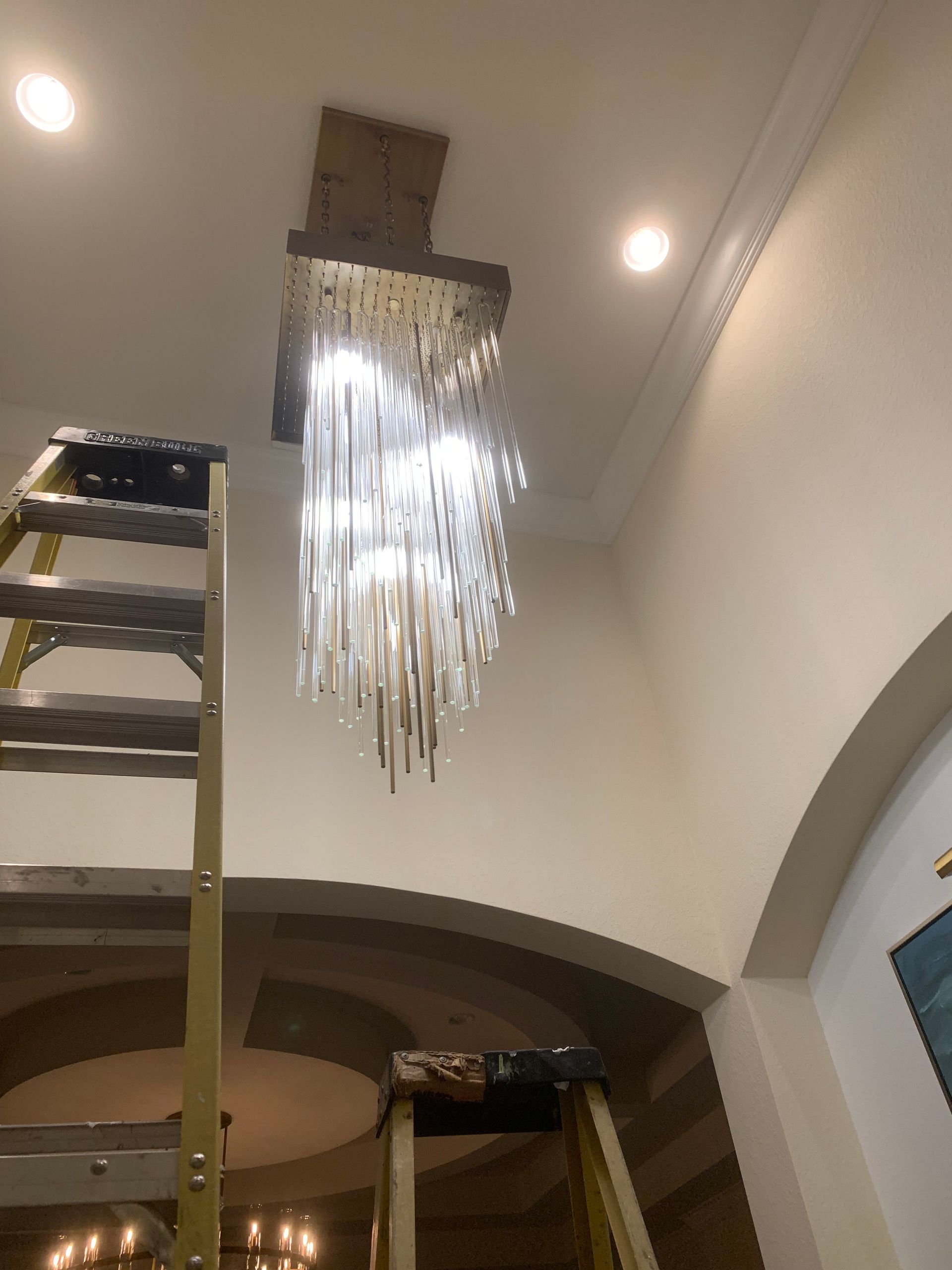 Chandelier being installed on a ceiling, with ladders in a bright interior hallway