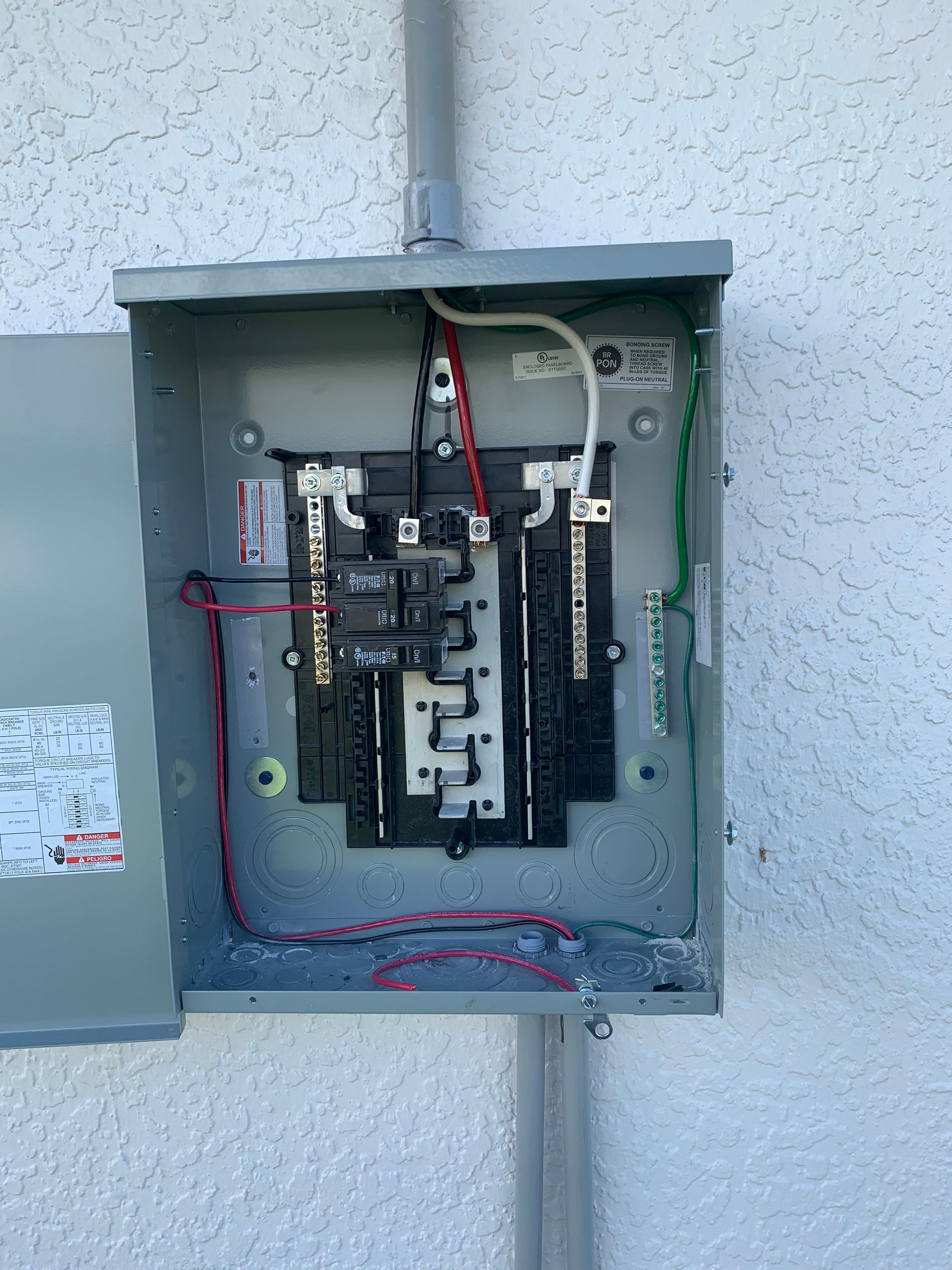 Open electrical panel mounted on an exterior wall with exposed wiring and breakers
