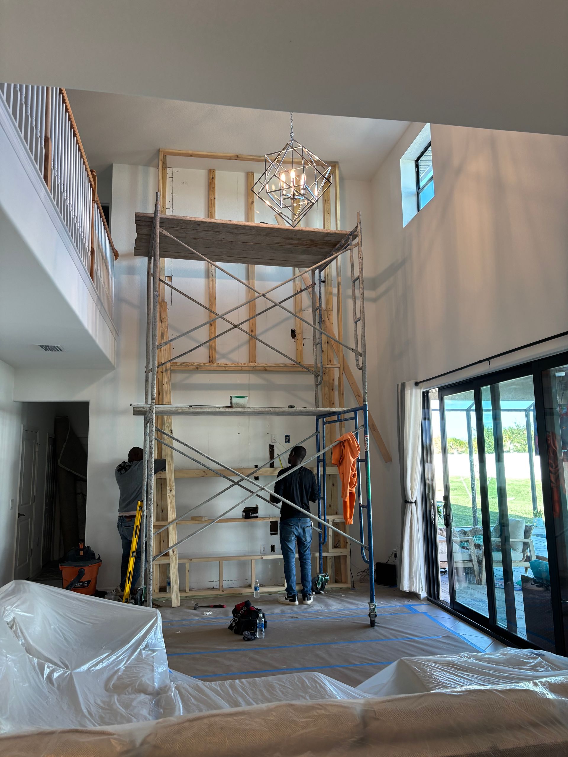 Tall living room with scaffolding, workers, and a hanging chandelier under a high ceiling.