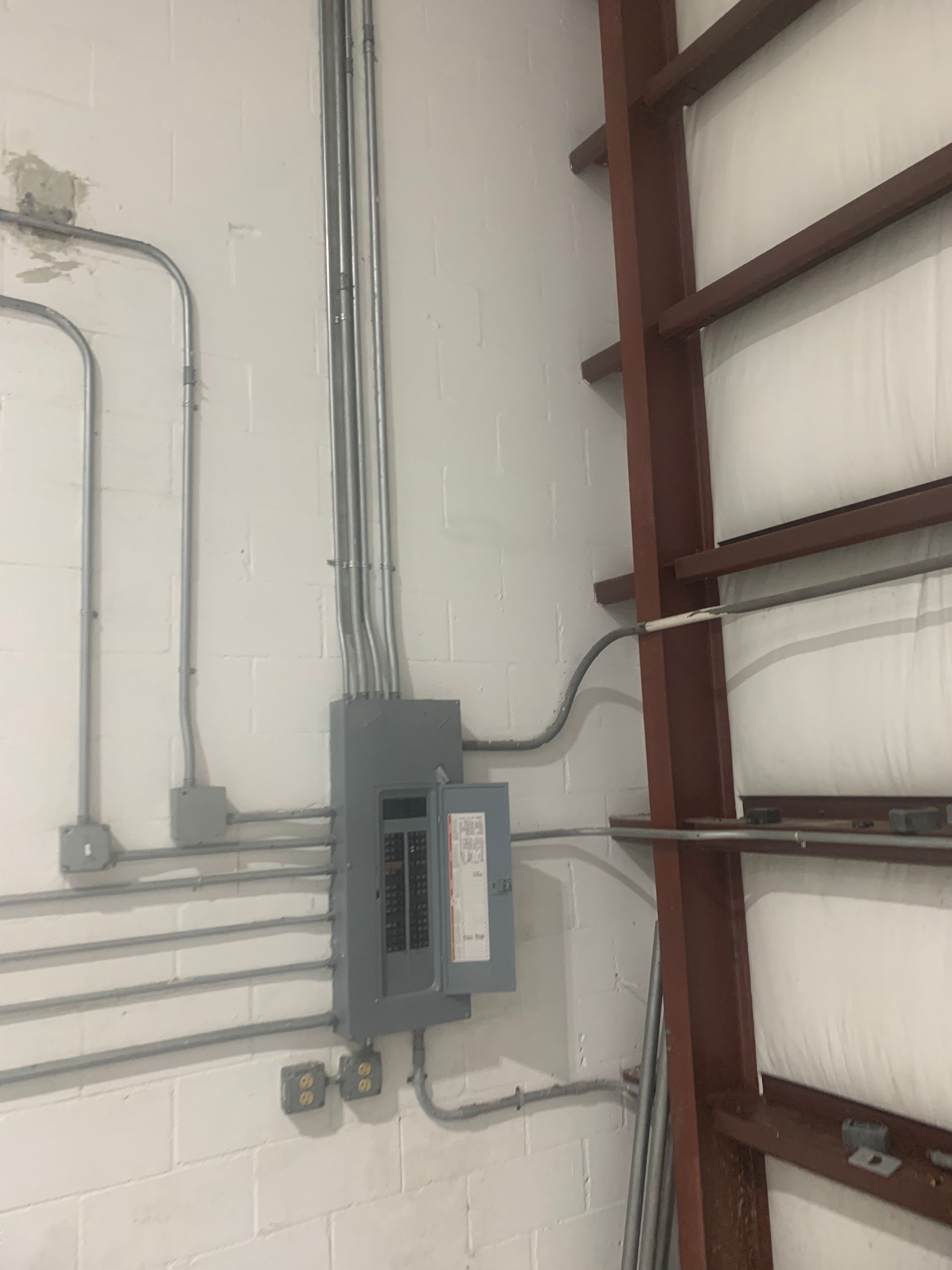 Gray electrical breaker panel with exposed conduit on a white cinderblock wall, next to red steel shelving.
