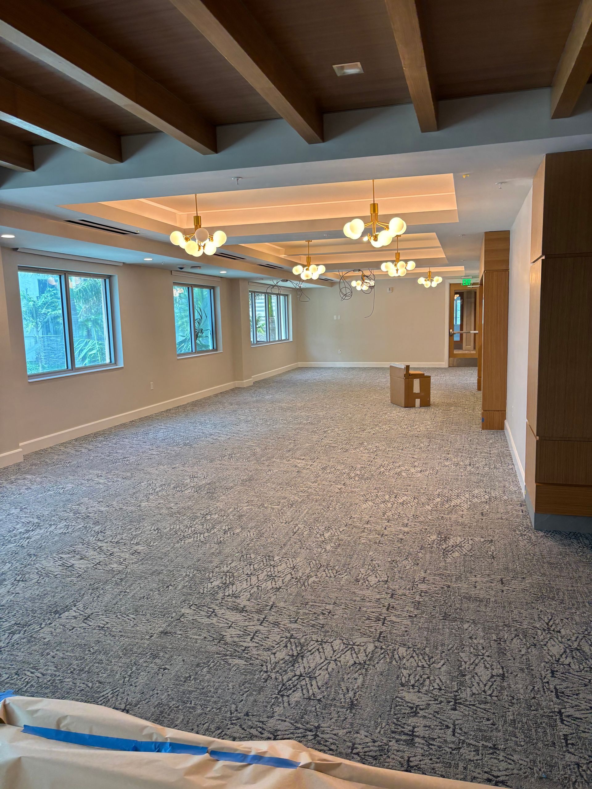 Empty carpeted banquet hall with wood-beam ceiling, chandeliers, and windows along the left wall.