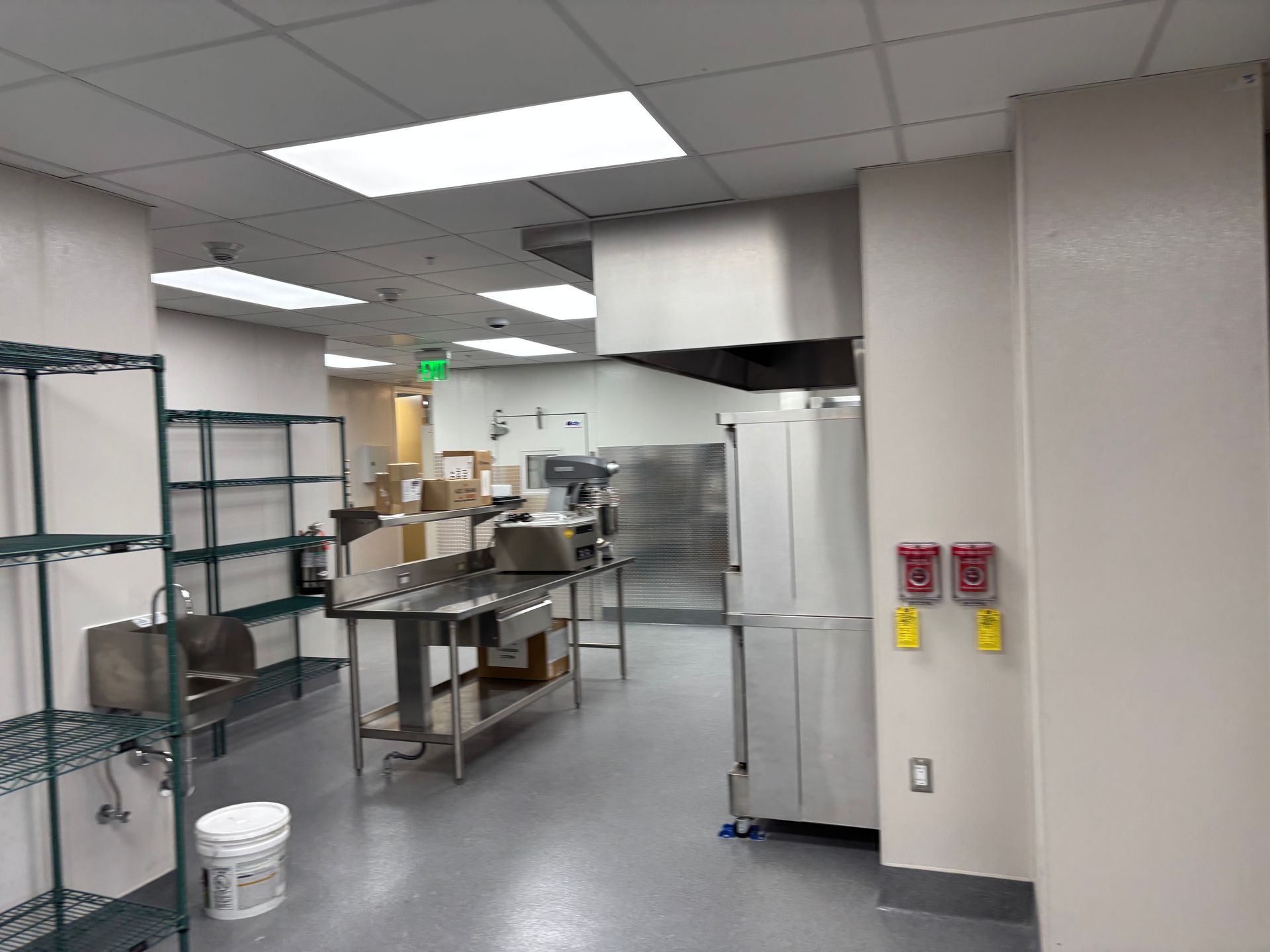 Commercial kitchen with stainless steel tables, shelves, and a white refrigerator under fluorescent lights