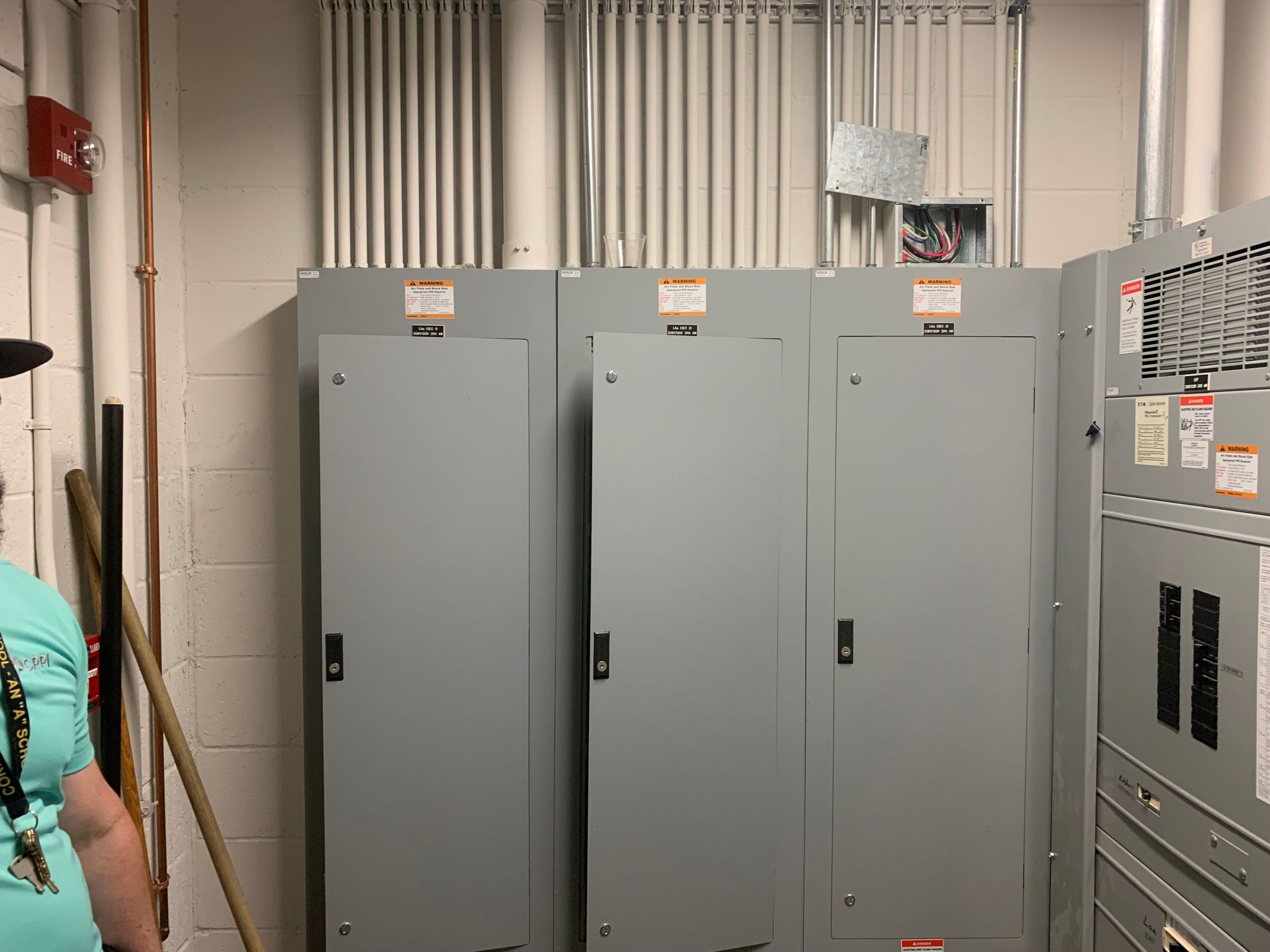Gray electrical control cabinets in an industrial room, with pipes and a partial person at the left edge.