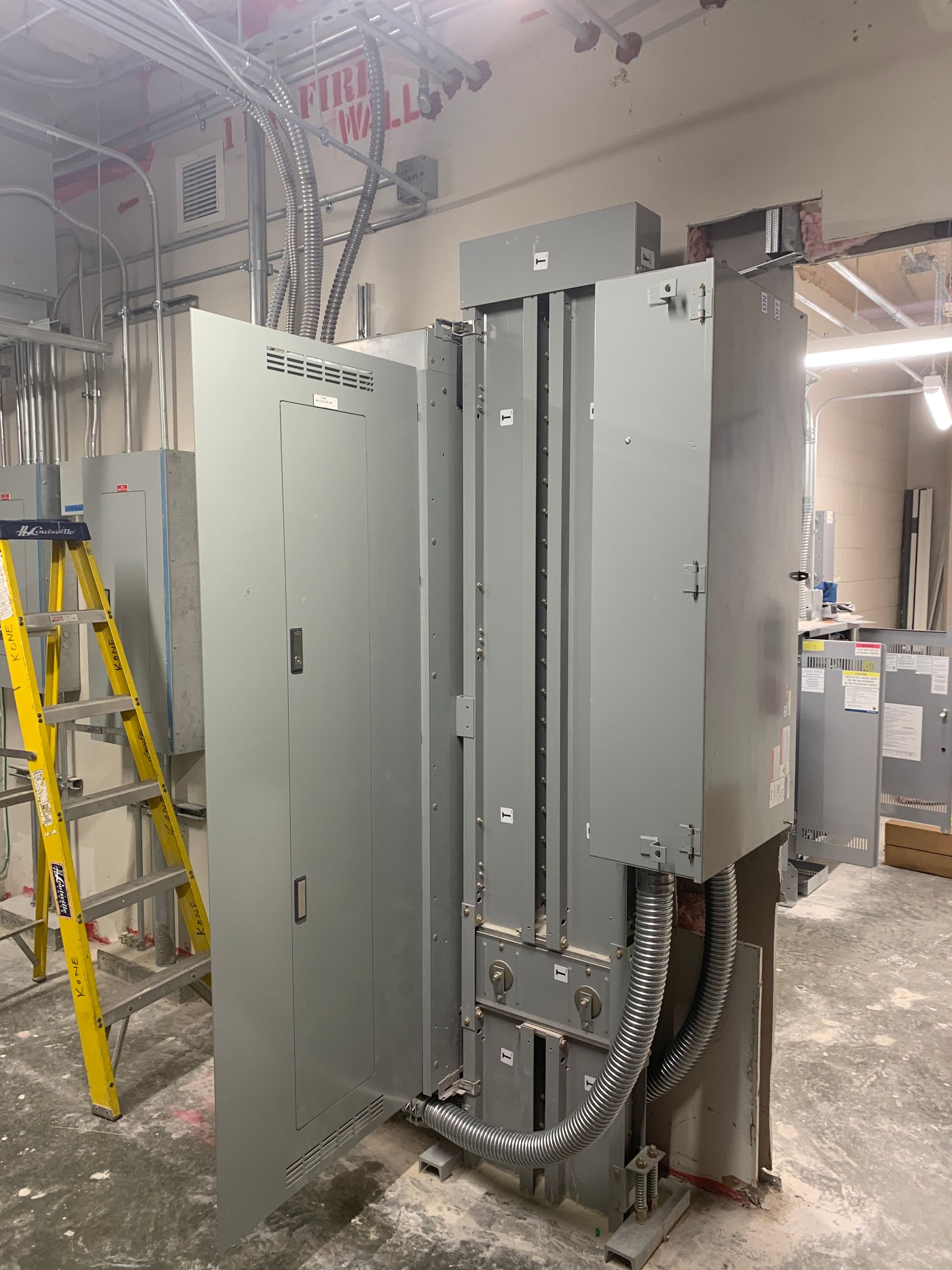 Open electrical control panels with wiring in a construction area, beside a yellow ladder