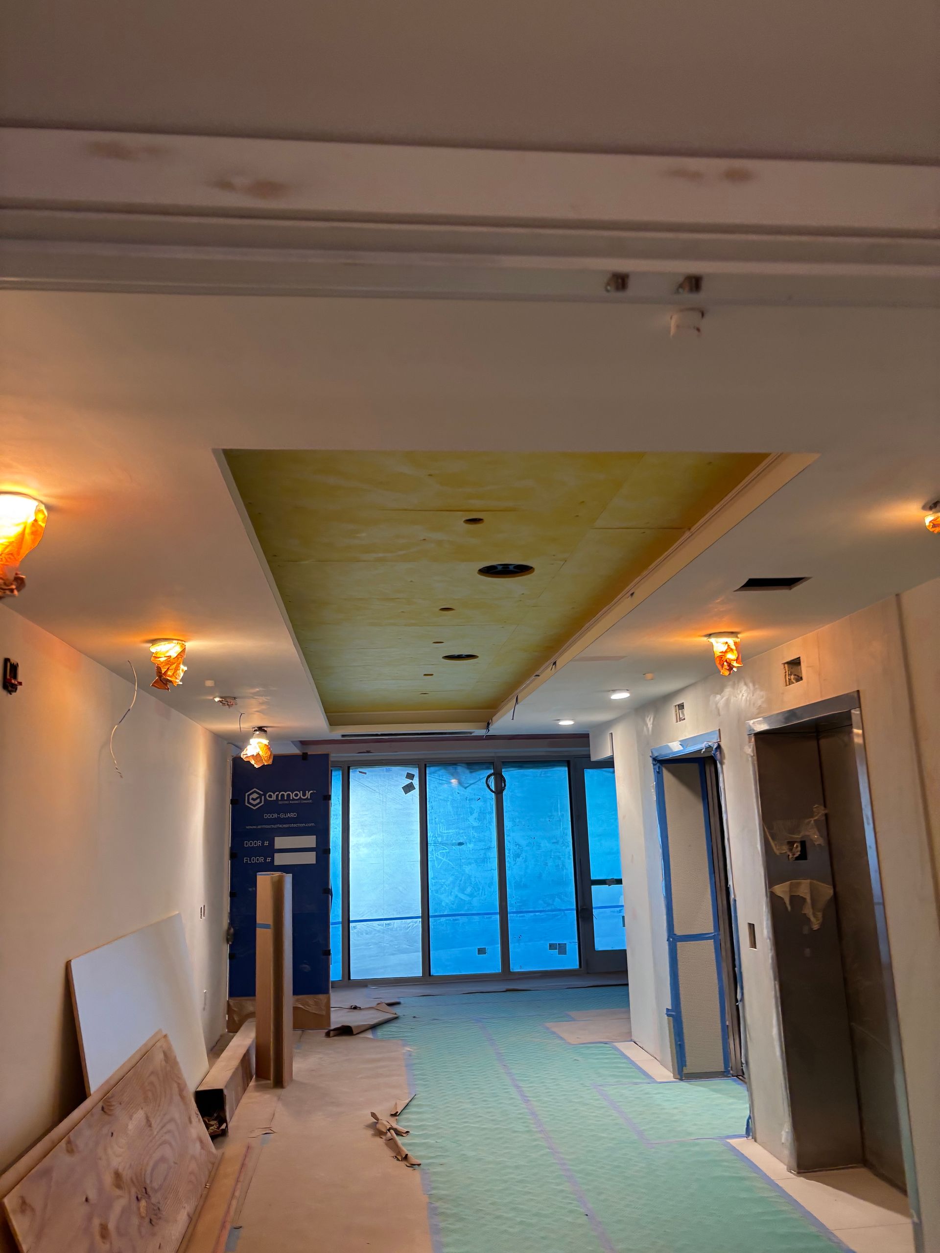 Narrow hallway under renovation with unfinished ceiling, exposed materials, and blue-covered doors at the end