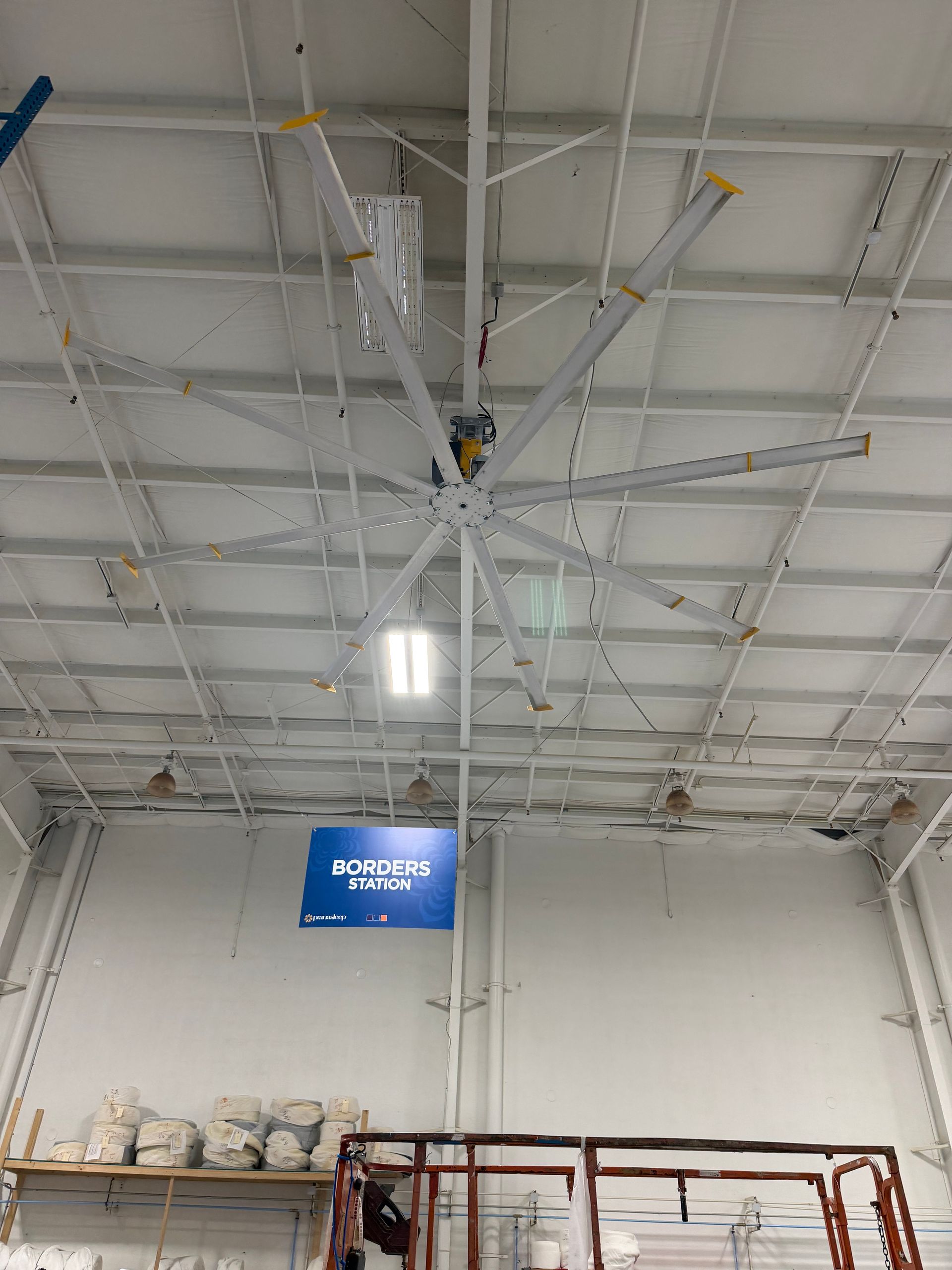 Warehouse ceiling with exposed beams, hanging fluorescent lights, and a blue sign above stacked shelving.