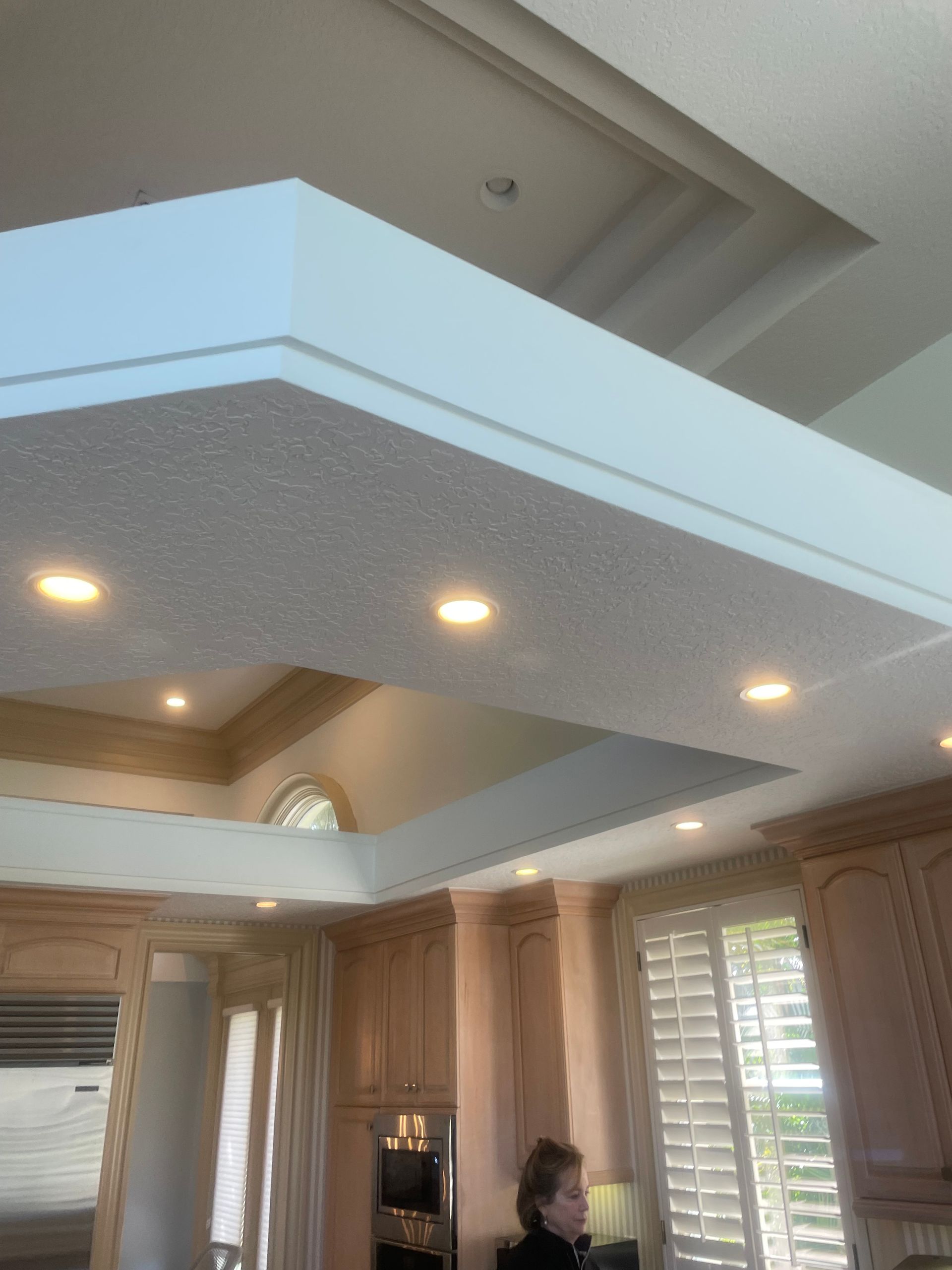 Modern kitchen with tray ceiling, recessed lights, wood cabinets, and a person standing near the counter.