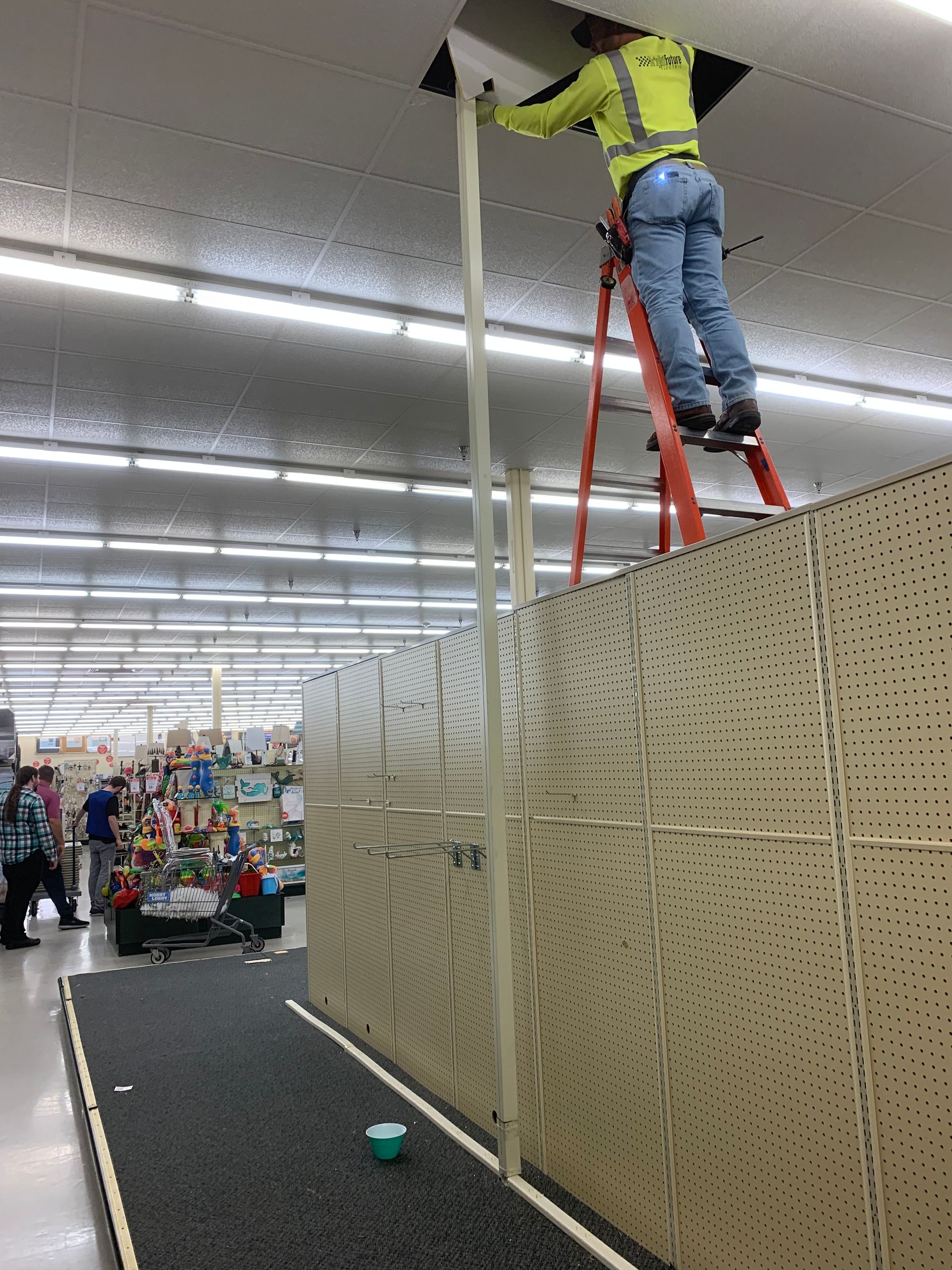 Worker in a neon shirt stands on a ladder fixing a ceiling display in a store aisle.