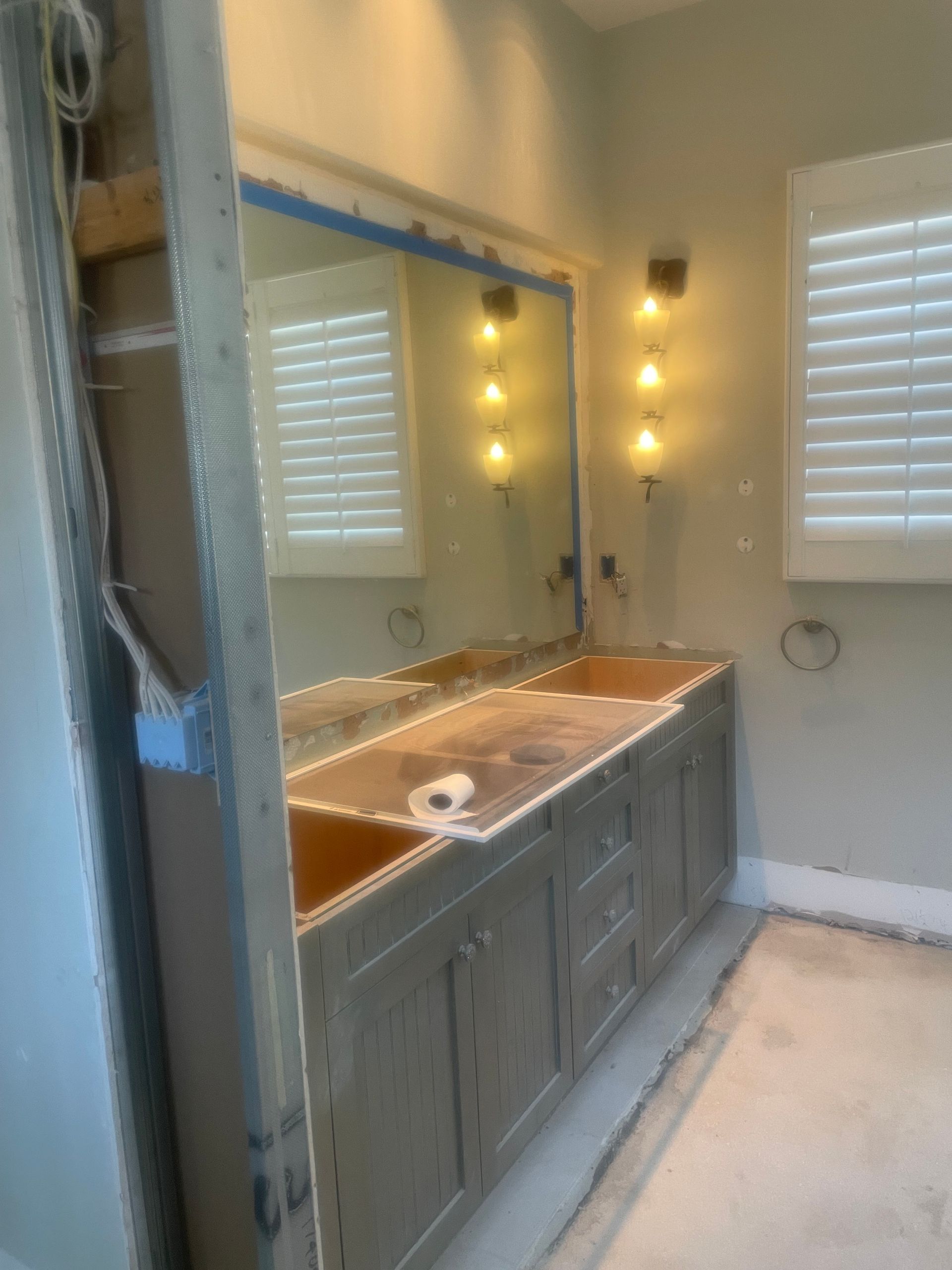 Bathroom vanity with large mirror, gray cabinets, and wall sconces beside window blinds.