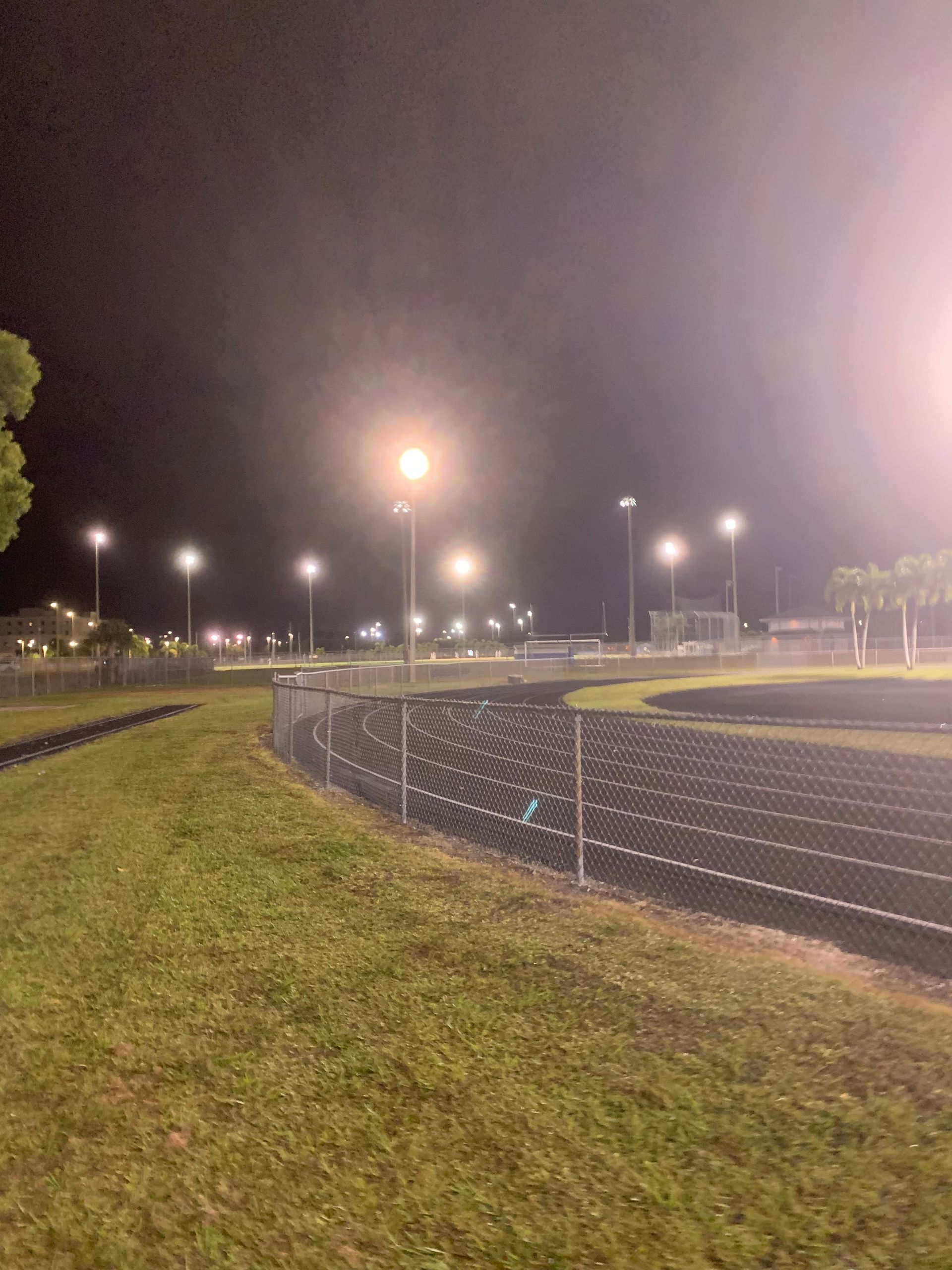 Nighttime running track with bright stadium lights, grassy infield, and a curved paved path alongside the oval track
