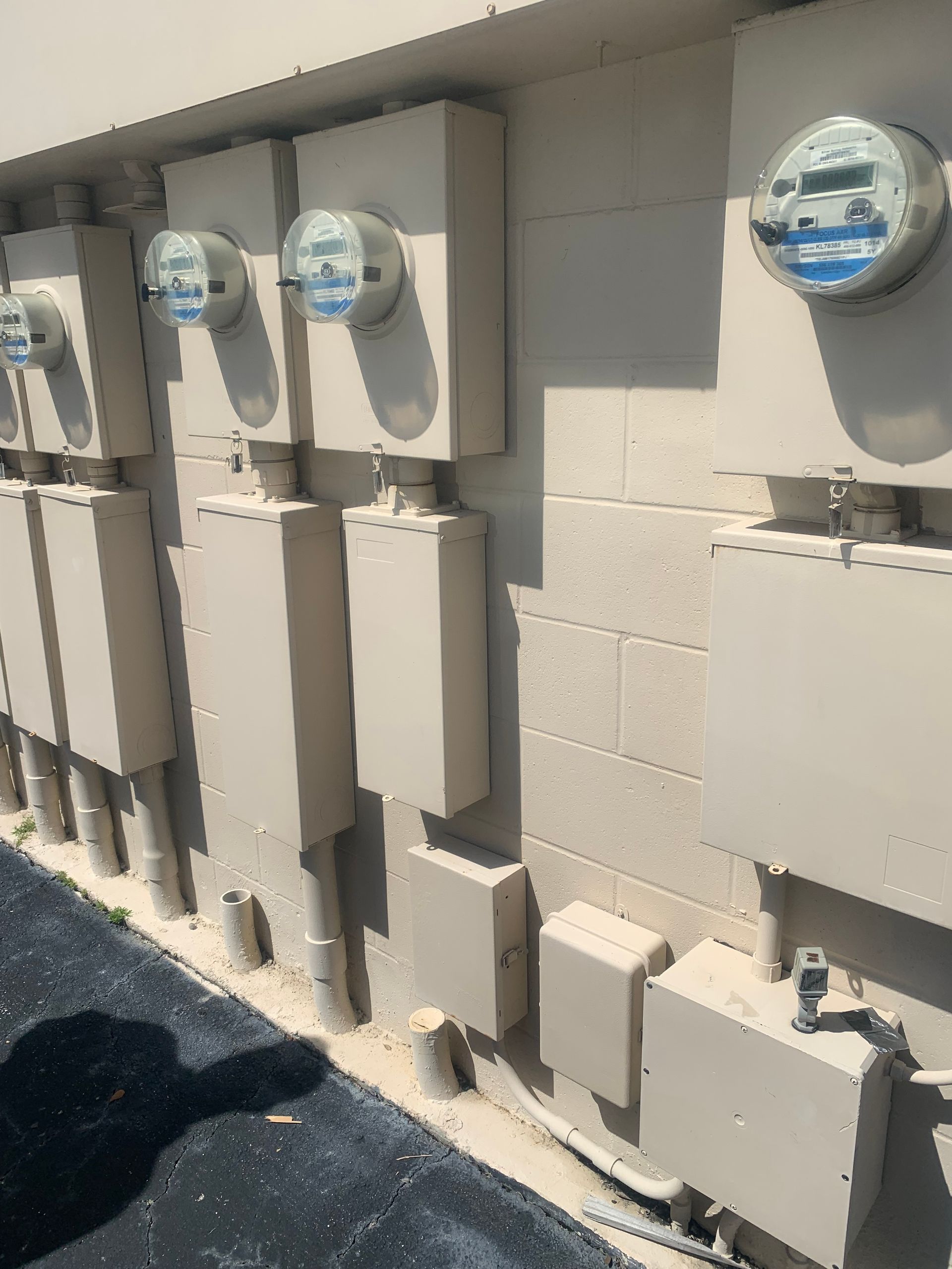 Row of electric meters and utility boxes mounted on a beige exterior wall in bright sunlight.