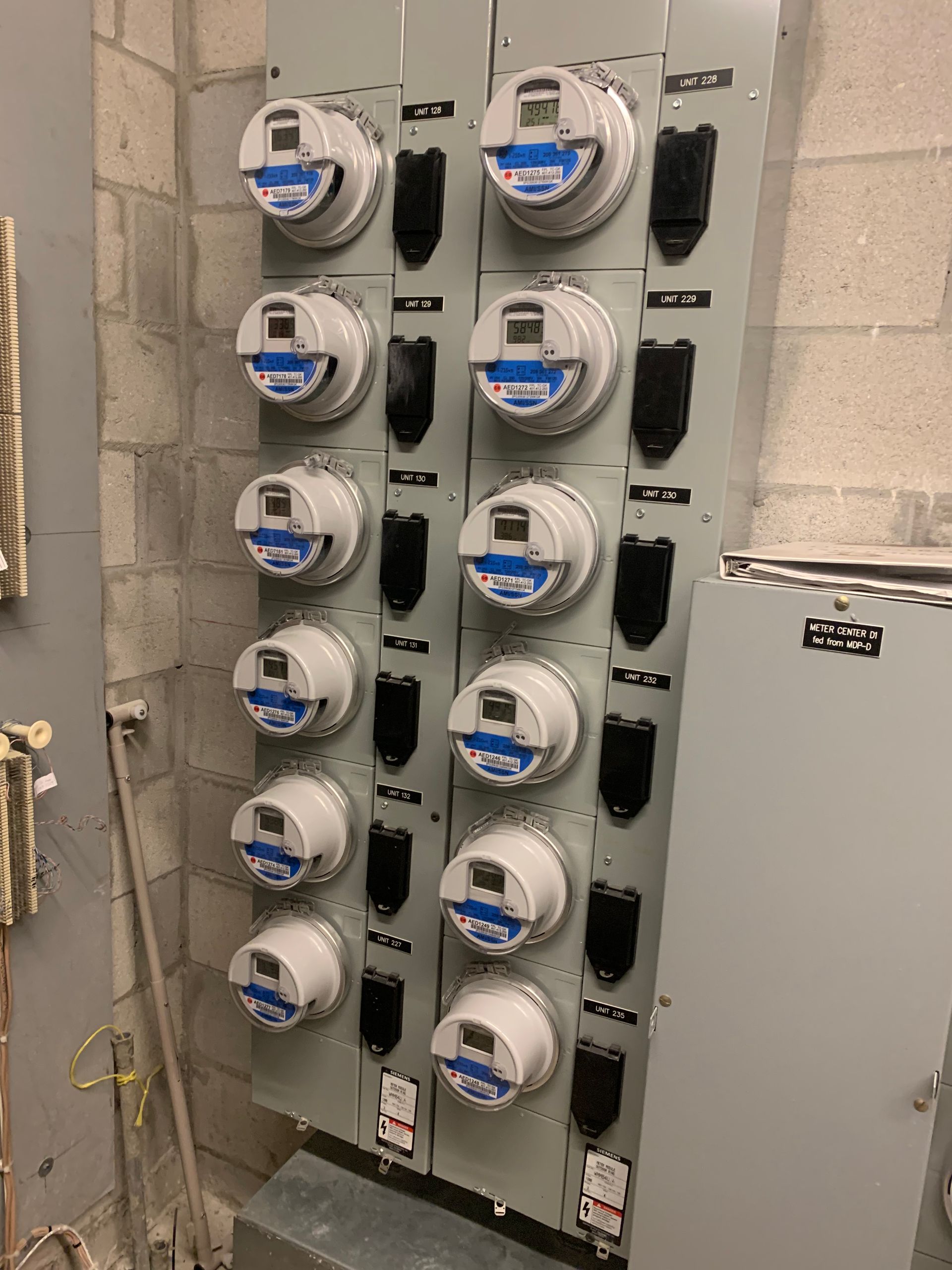 Electric utility meters mounted in a row on a wall next to electrical panels.