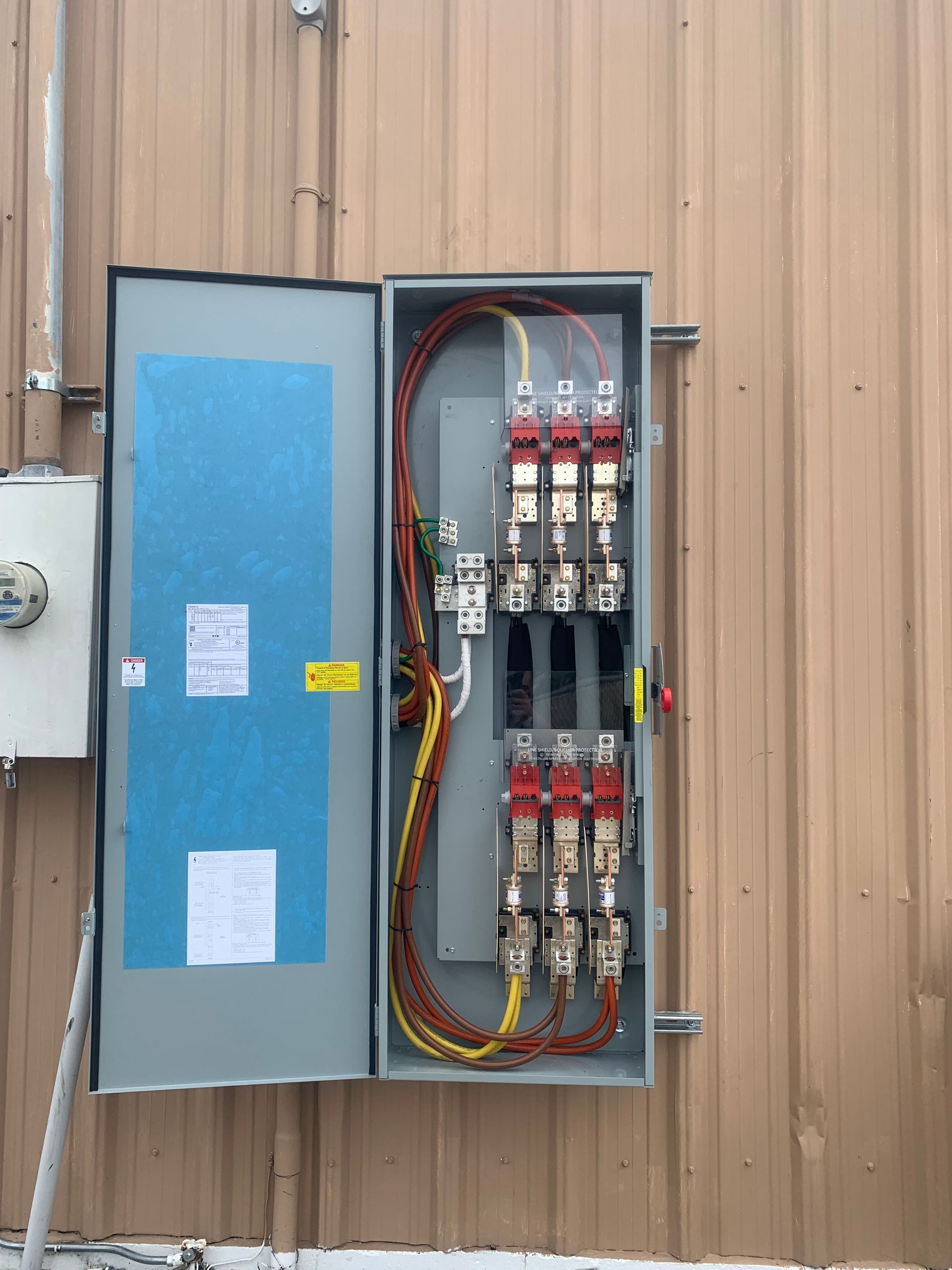 Open electrical breaker panel on tan metal building wall with exposed wiring and red switches