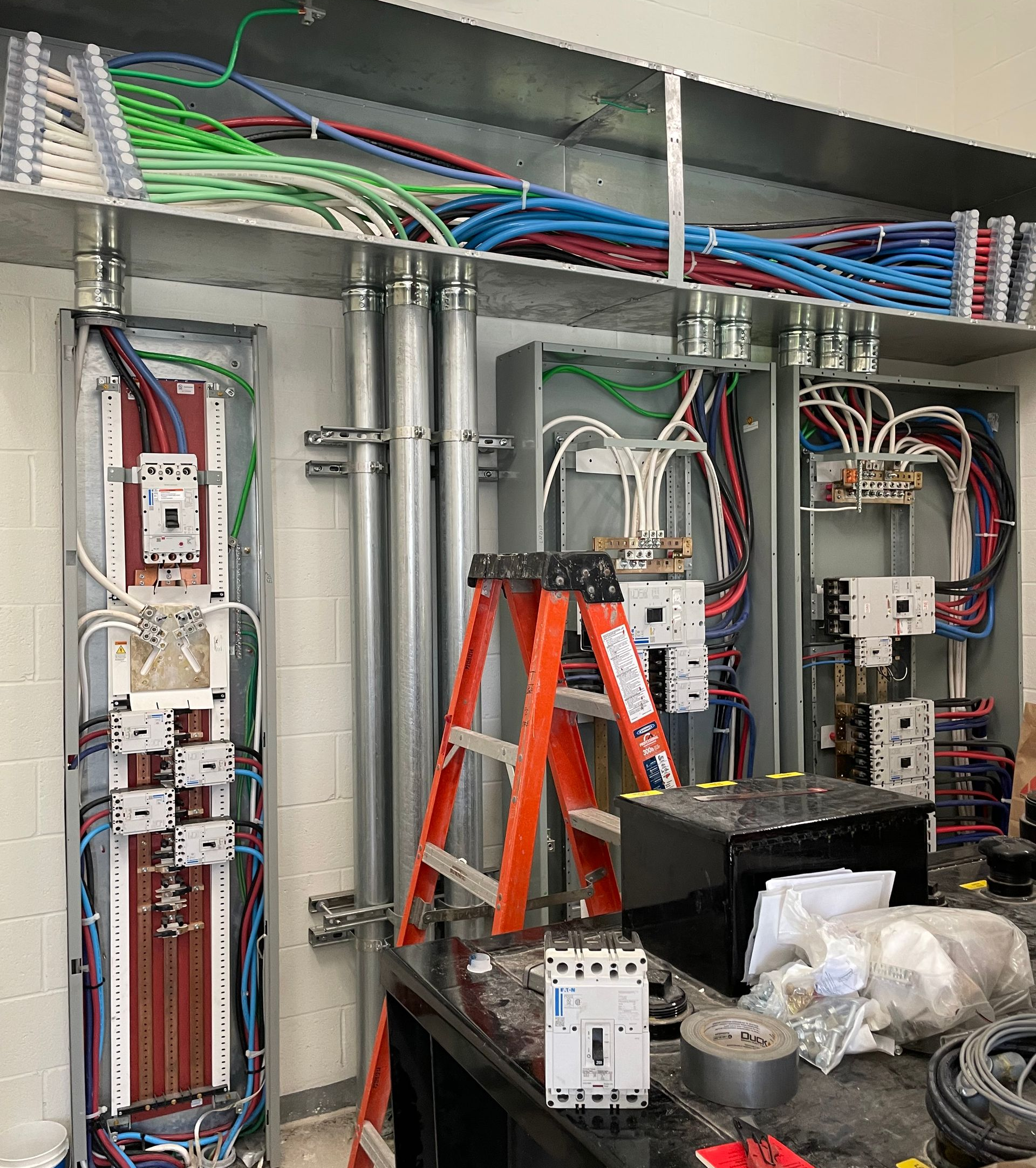 Open electrical control cabinet with wiring, breakers, and orange ladder inside a utility room