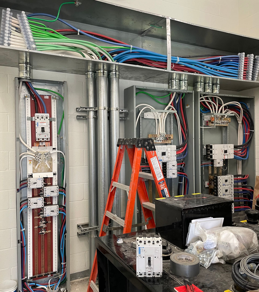 Open electrical control cabinet with wiring, breakers, and orange ladder inside a utility room