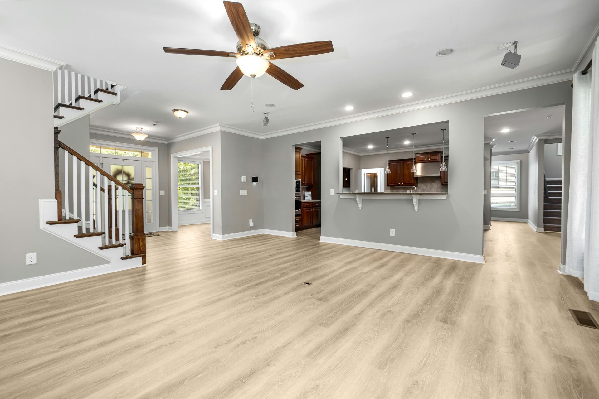 Spacious open-plan living room with light wood floors, gray walls, ceiling fan, staircase, and kitchen pass-through.