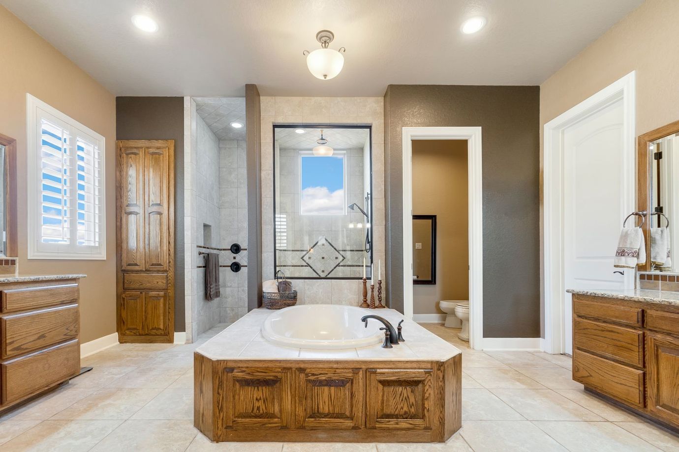 Bright bathroom with a freestanding tub, glass shower, wood vanities, and beige tile floors.