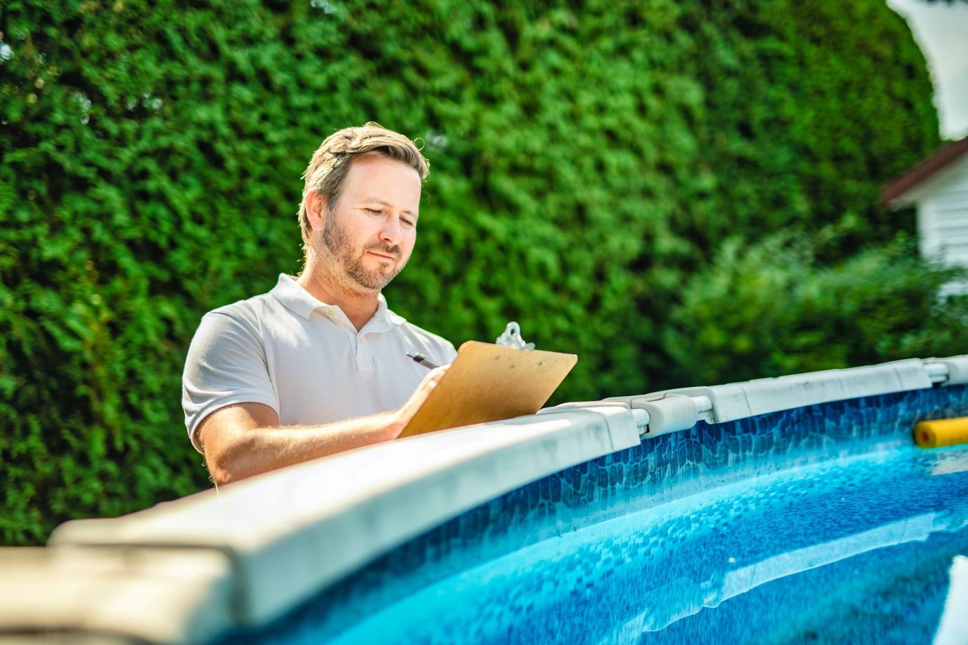 Man holding a clipboard beside a blue swimming pool in a sunny backyard