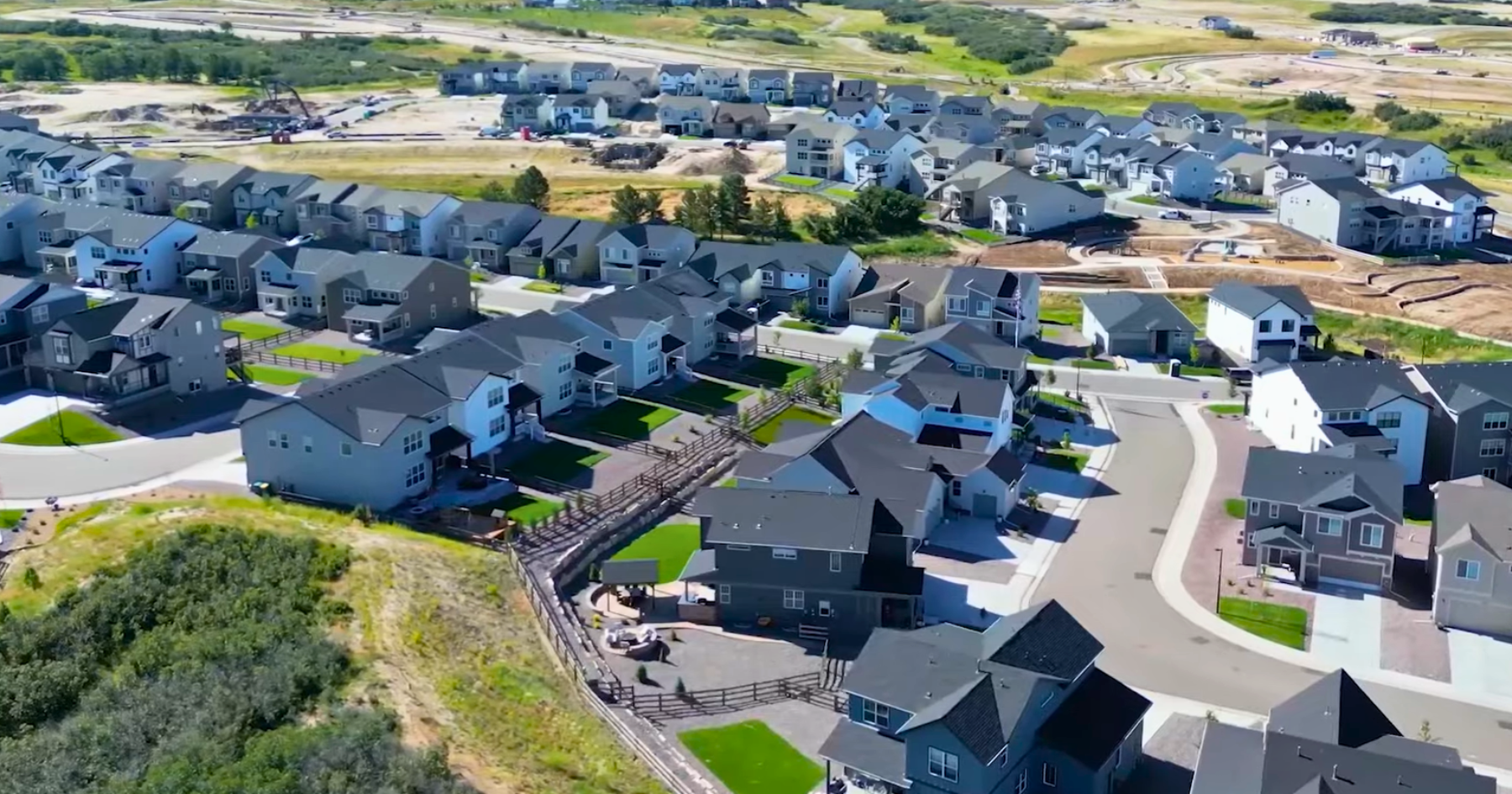 Aerial view of a suburban neighborhood featuring numerous detached houses with gray roofs and yards near a grassy hill.