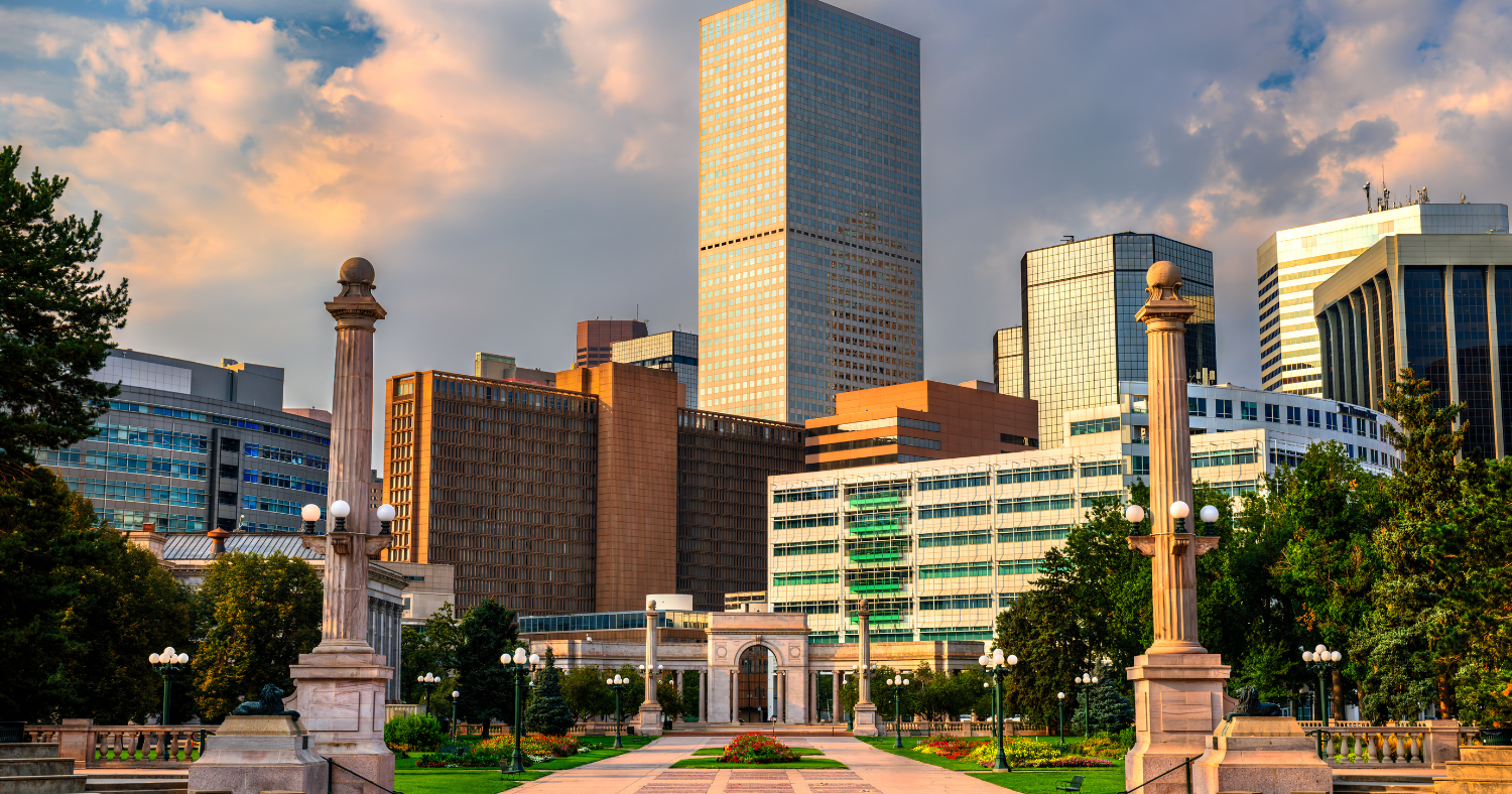 Civic Center Park in Denver with its tall stone pillars, landscaped gardens, and downtown skyscrapers under a cloudy sky.
