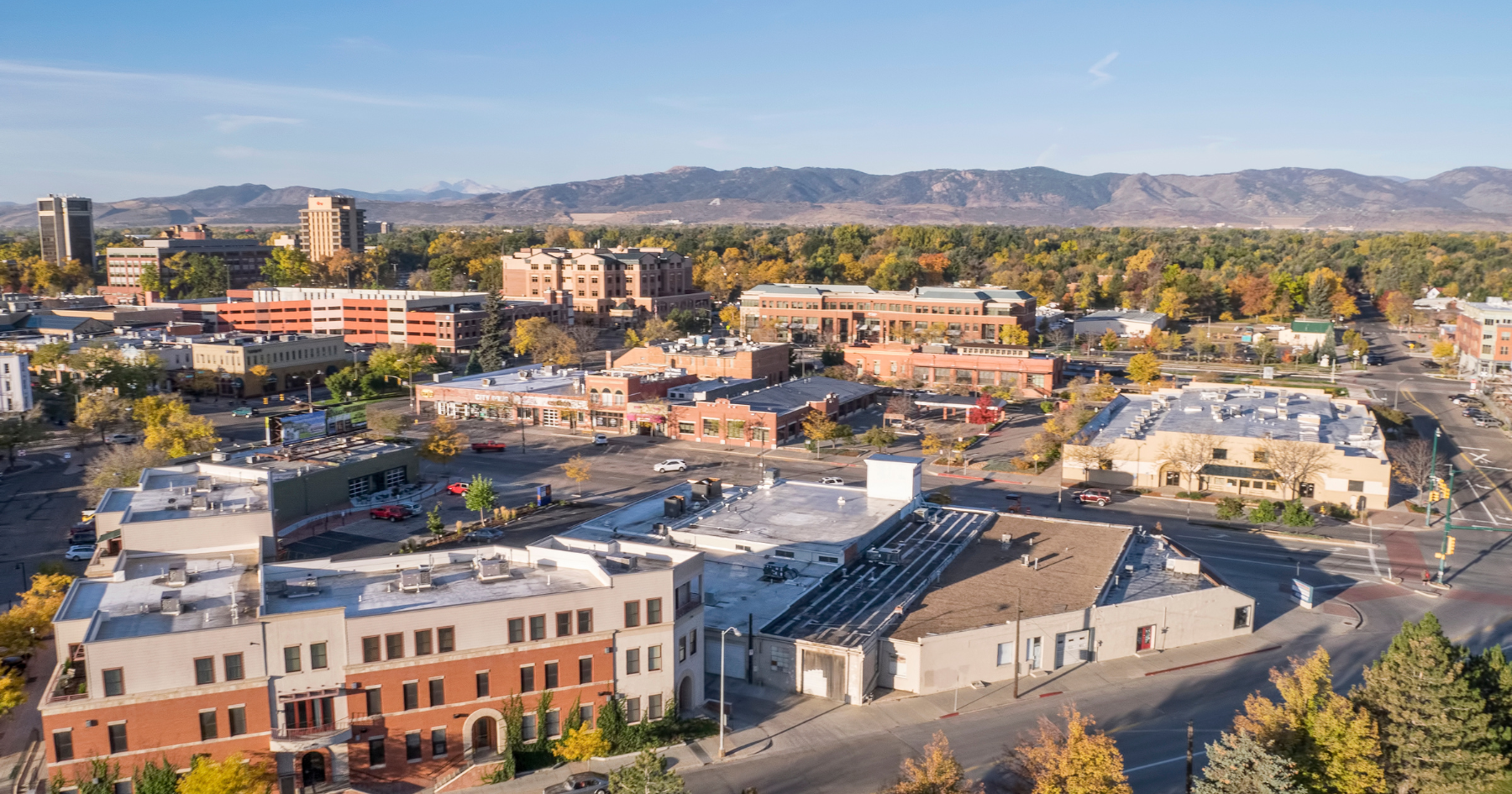 Aerial view of a town with low-rise buildings, autumnal trees, and a mountain range in the distance under a blue sky.