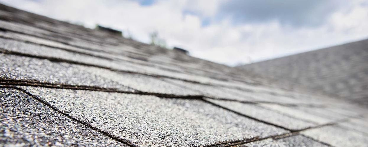Close-up perspective of gray asphalt roof shingles slanting toward a ridge against a cloudy sky.
