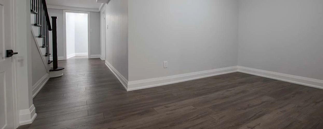 An empty room with gray walls, dark wood flooring, white baseboards, and a staircase leading to an open doorway.