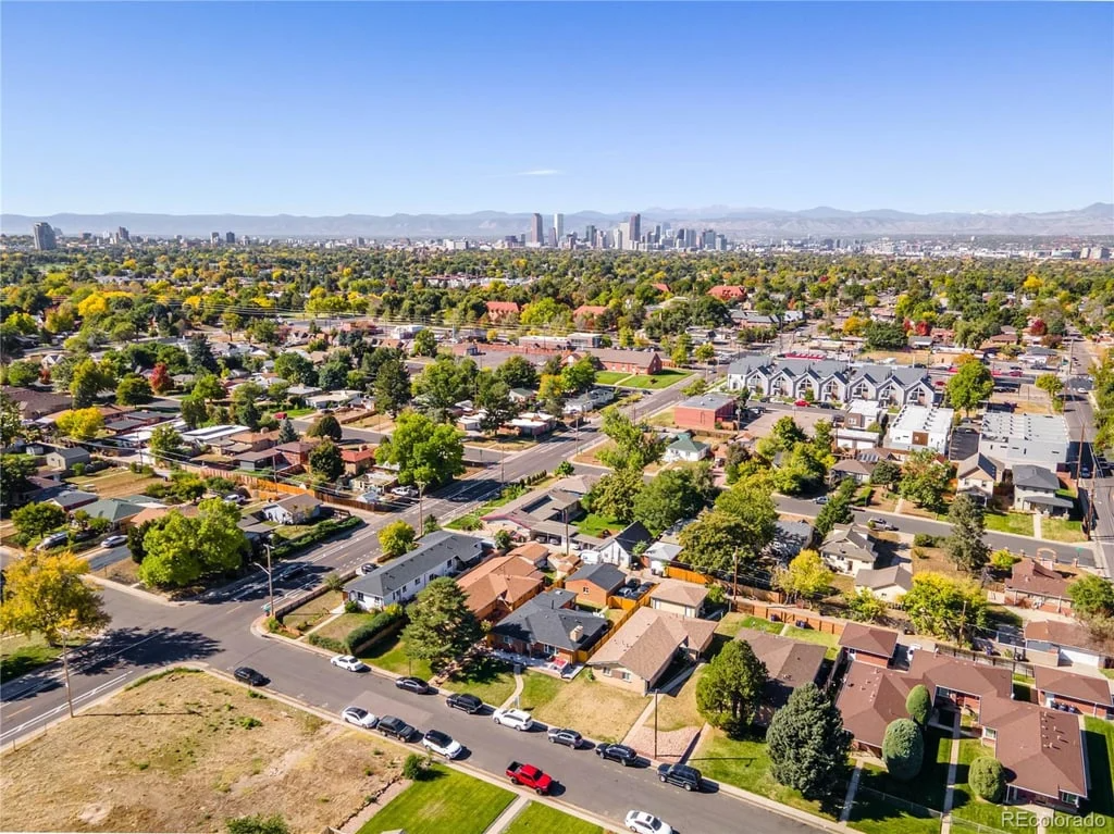 Aerial view of a residential neighborhood with tree-lined streets and a distant city skyline under a clear blue sky.