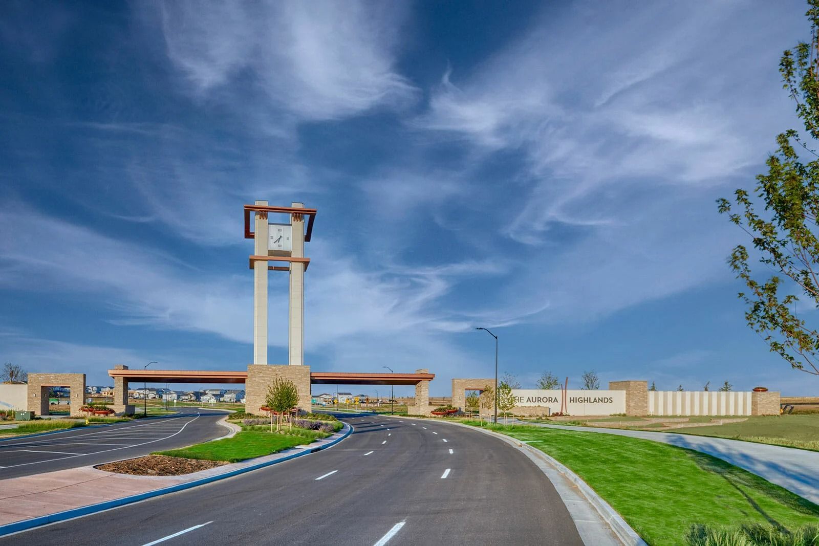 A modern entrance gate with a clock tower stands over a paved road under a bright blue sky with wispy clouds.