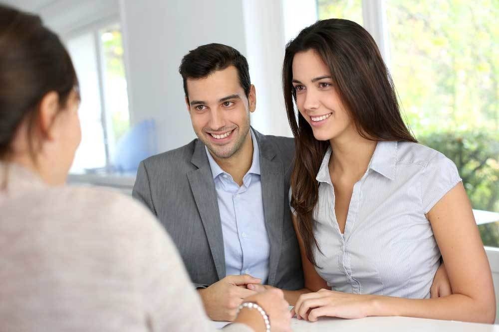 A smiling couple in professional attire sits at a table, looking attentively toward a person across from them.