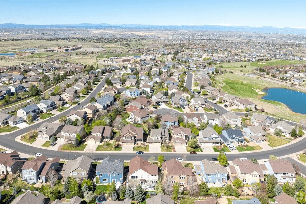 Aerial view of a suburban neighborhood with many single-family homes, paved roads, and a nearby pond under a clear sky.