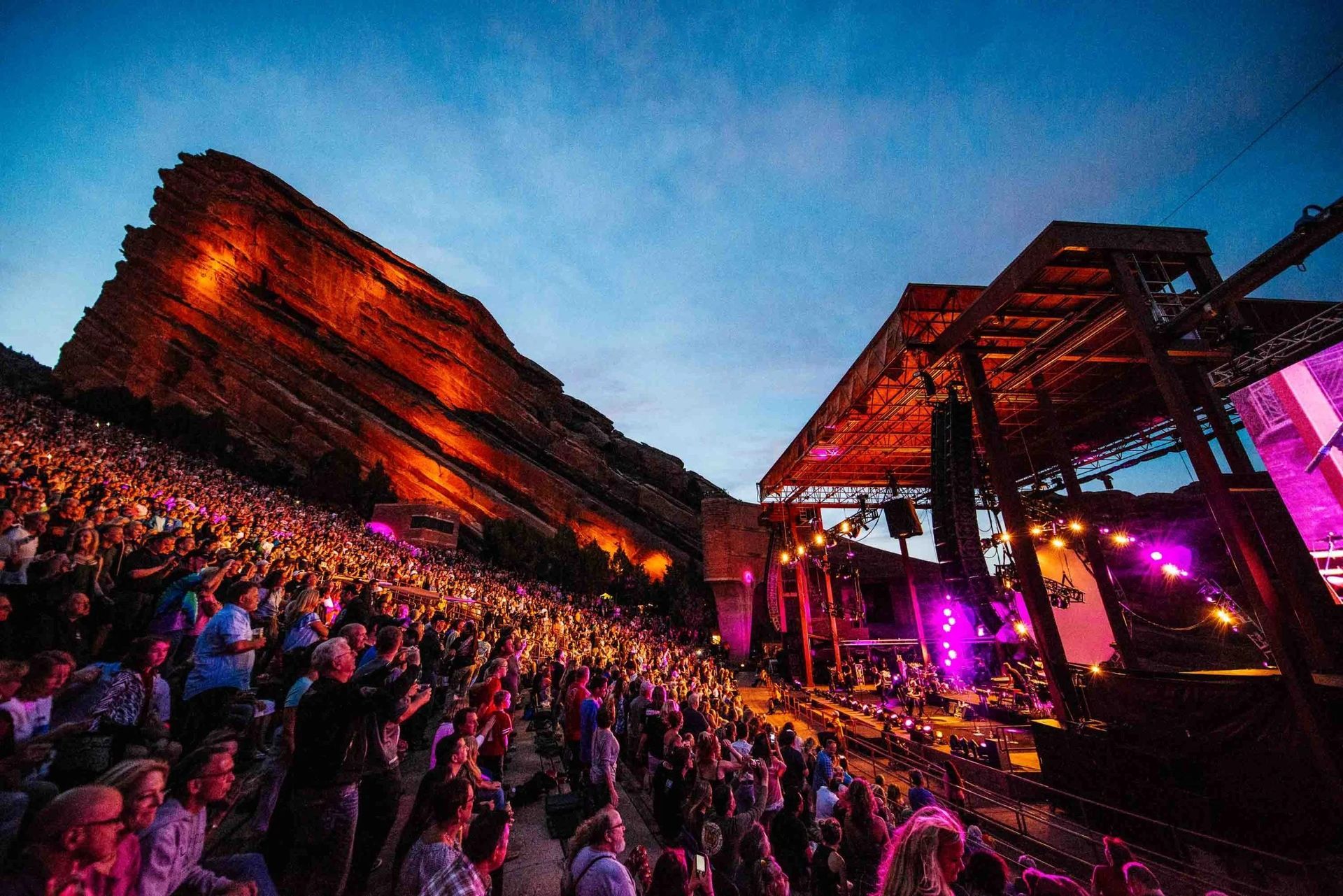 A large, energetic crowd watches a concert at sunset in the famous Red Rocks Amphitheatre, with dramatic rock walls.