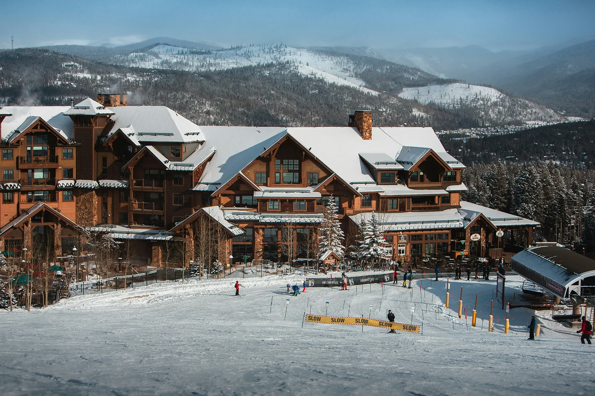 A large, multi-story wooden mountain lodge sits at the base of a snowy ski slope under a clear blue sky.
